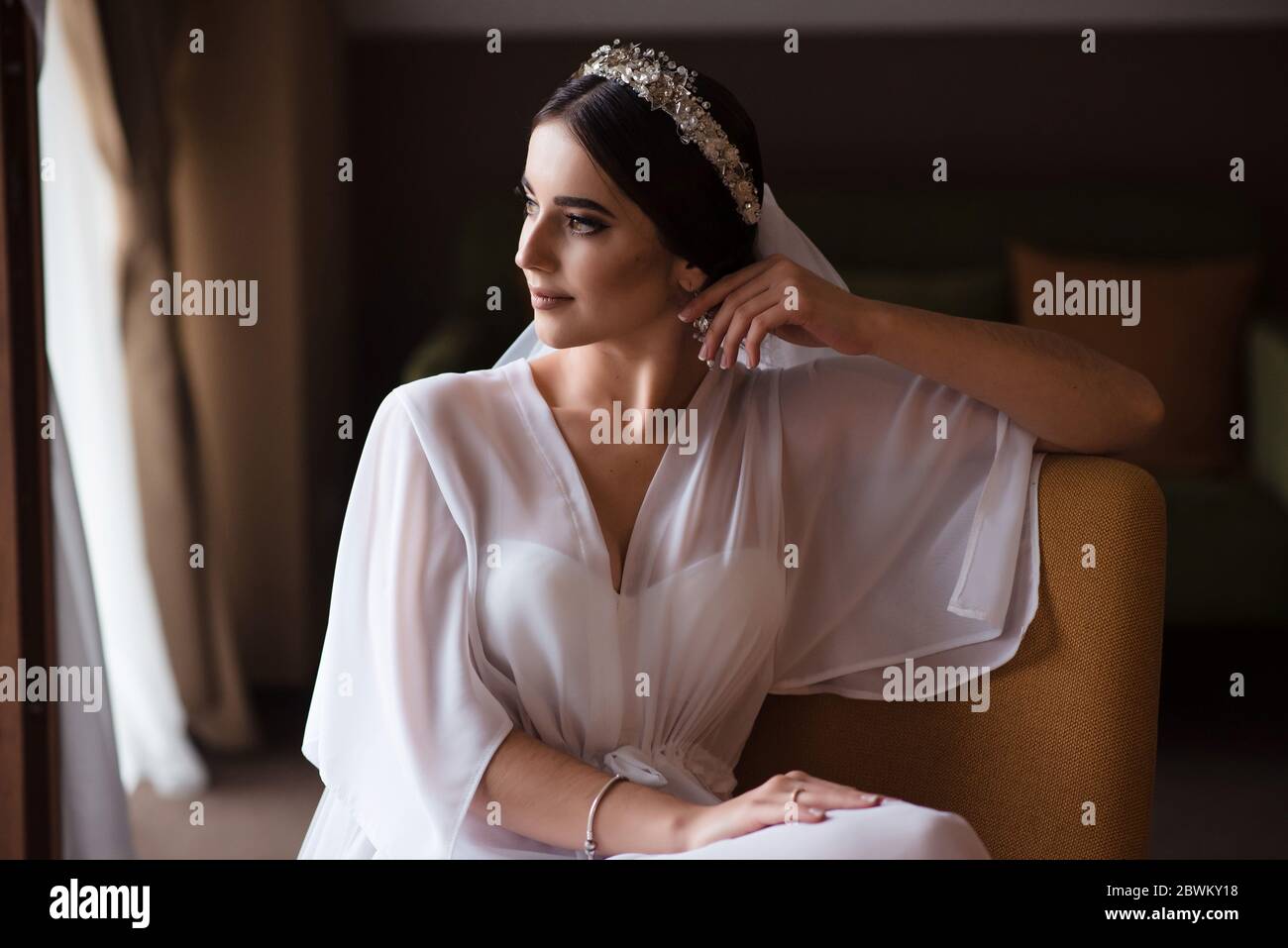 Novia de mañana en una habitación de hotel. Novia morena con maquillaje y  una tiara en su cabello se sienta en una silla y mira por la ventana  Fotografía de stock -