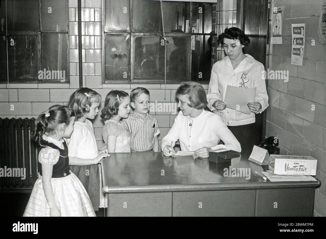 Educación en los EE.UU. En la década de 1950 – aquí en una oficina de azulejos una enfermera o visitante de salud está viendo y cuestionando a cuatro niños en la escuela primaria. Está utilizando un sistema de índice de tarjetas. En su escritorio hay un hisopo en un tubo de vidrio y una caja de pañuelos de papel. Hay carteles relacionados con la salud en la pared. Foto de stock