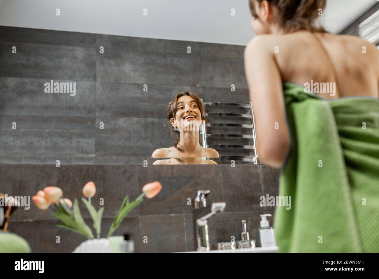 Joven y hermosa mujer cuidándose de sí misma, mirando en el gran espejo del baño en casa Foto de stock