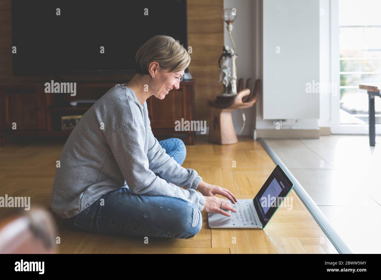 Mujer madura trabajando desde casa, usando un portátil, sentada de patas cruzadas en el suelo Foto de stock
