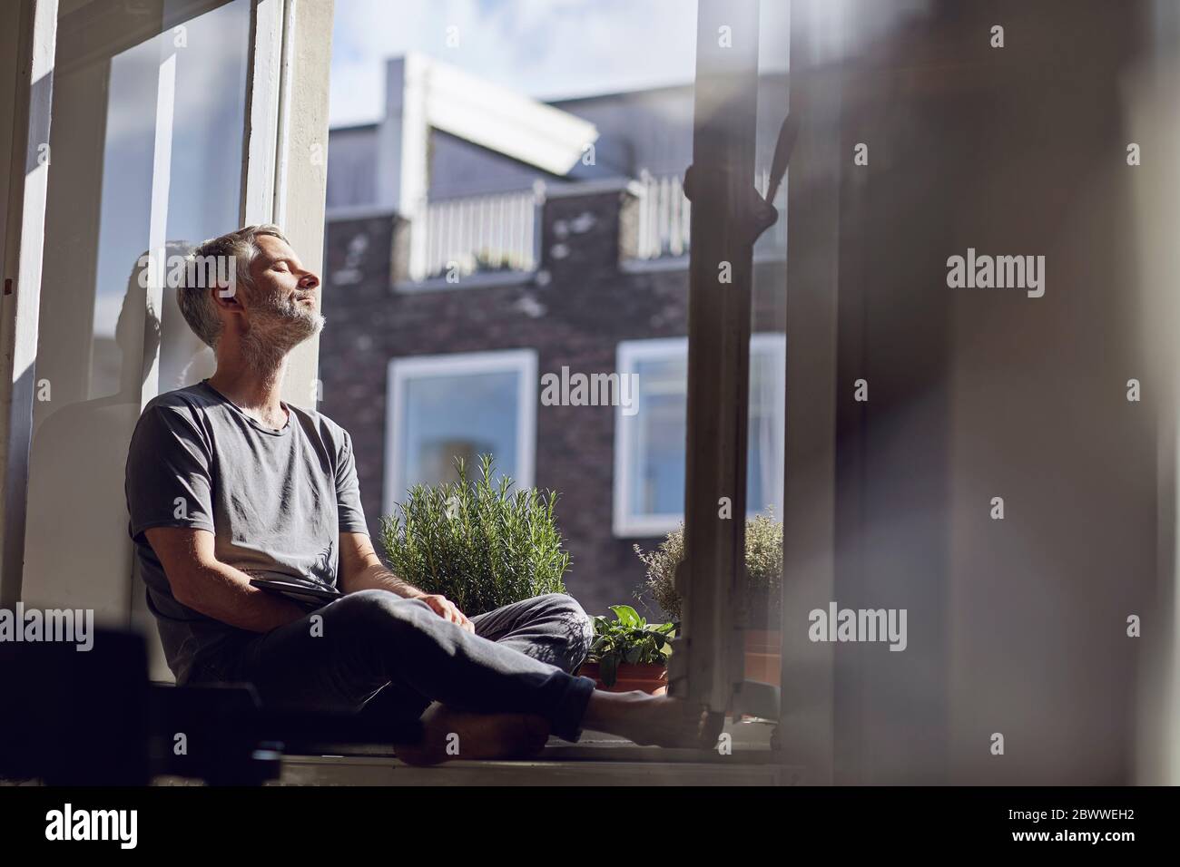 Hombre maduro sentado en la ventana de casa con los ojos cerrados Foto de stock