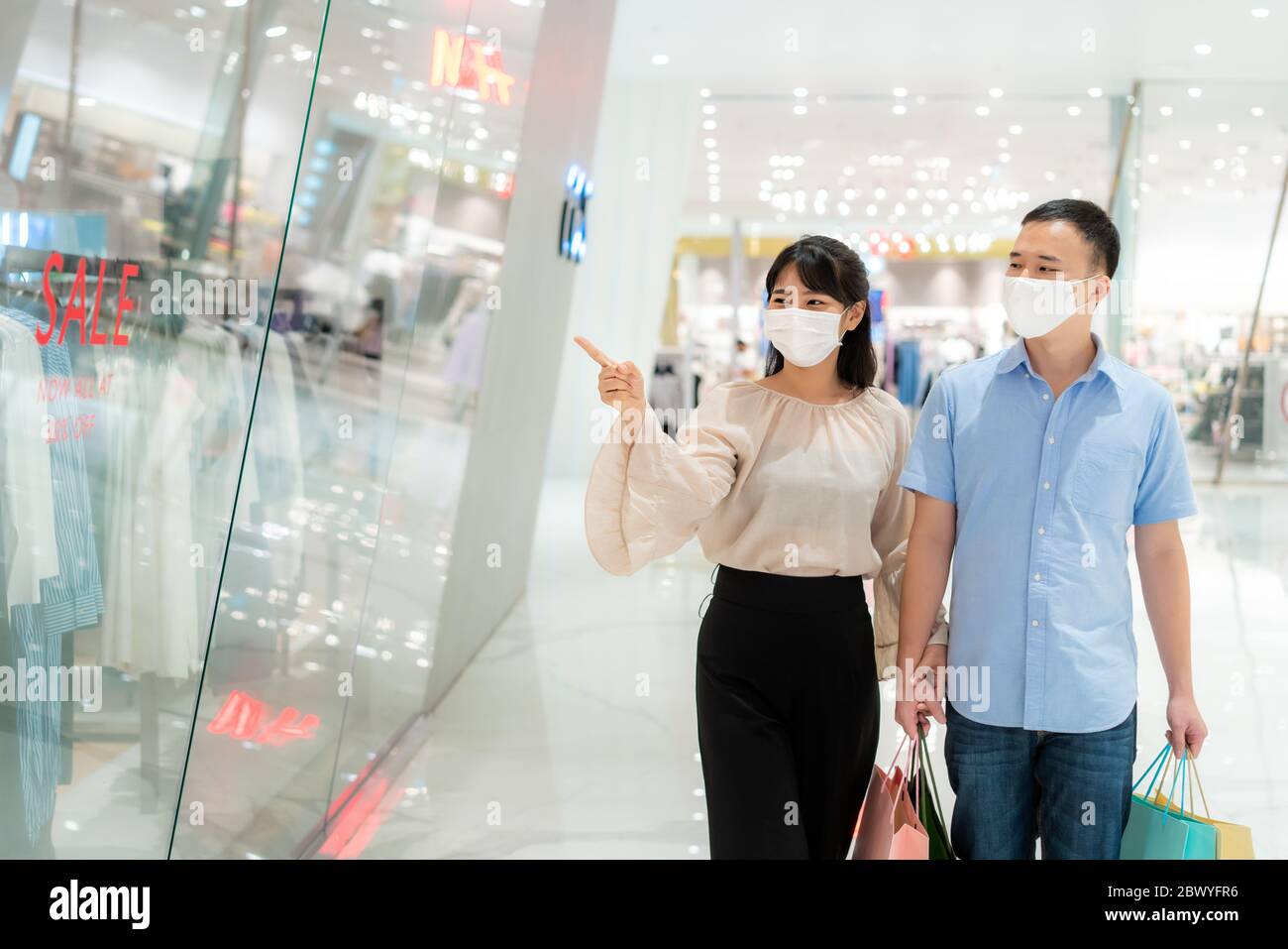 Pareja asiática usando máscara sobre su cara mirando nueva colección de ropa en el centro comercial con bolsa de compras para la atención médica y la prevención de coronavir Foto de stock