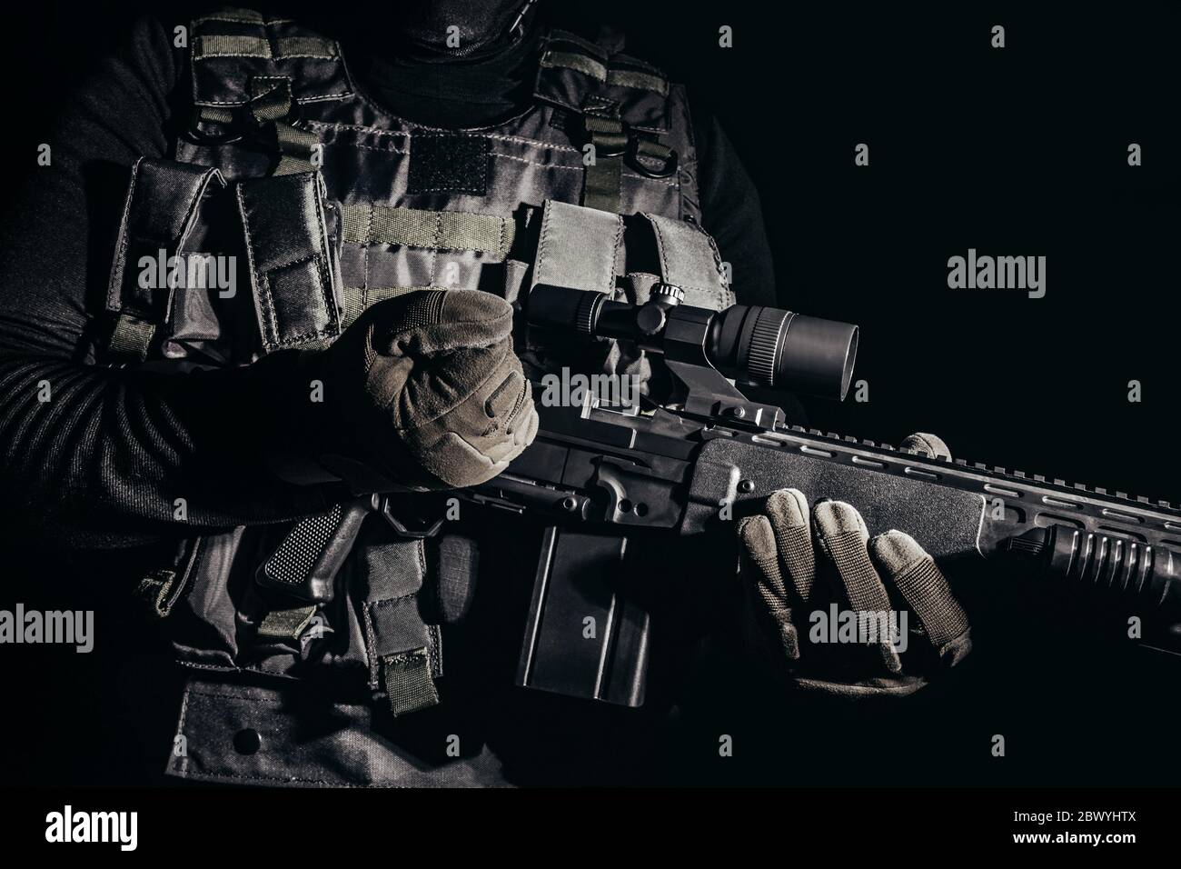 Foto de un soldado con guantes tácticos y chaleco de armadura de pie y recarga de rifle de francotirador sobre fondo negro. Foto de stock