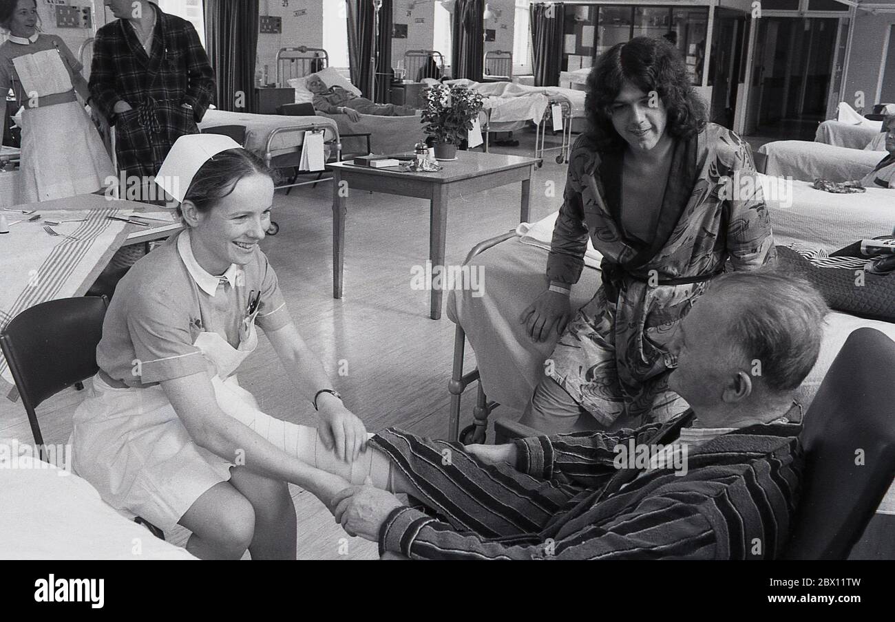 Años 70, histórica, una joven enfermera sentada en una silla con la pierna de un anciano paciente en su regazo, vigilada por un joven paciente masculino, mientras charlan juntos en un hospital en Lewisham, Londres del Sur, Inglaterra, Reino Unido. Foto de stock