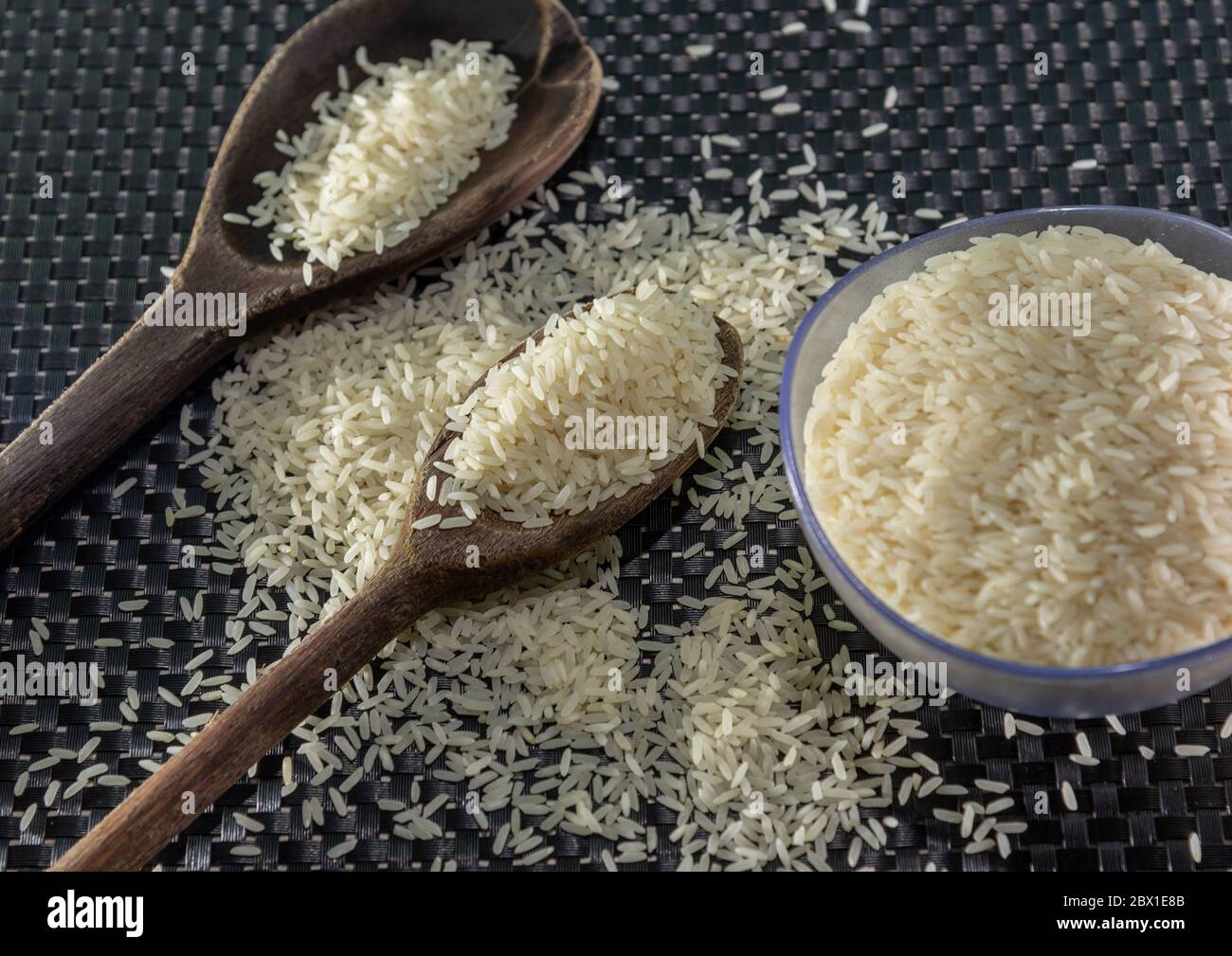 Arroz pelado y procesado de grano sobre fondo oscuro. Cereales de arroz y ornamentos de cocina. Granos semi-procesados en una olla de plástico y cucharas de madera. Foo Foto de stock