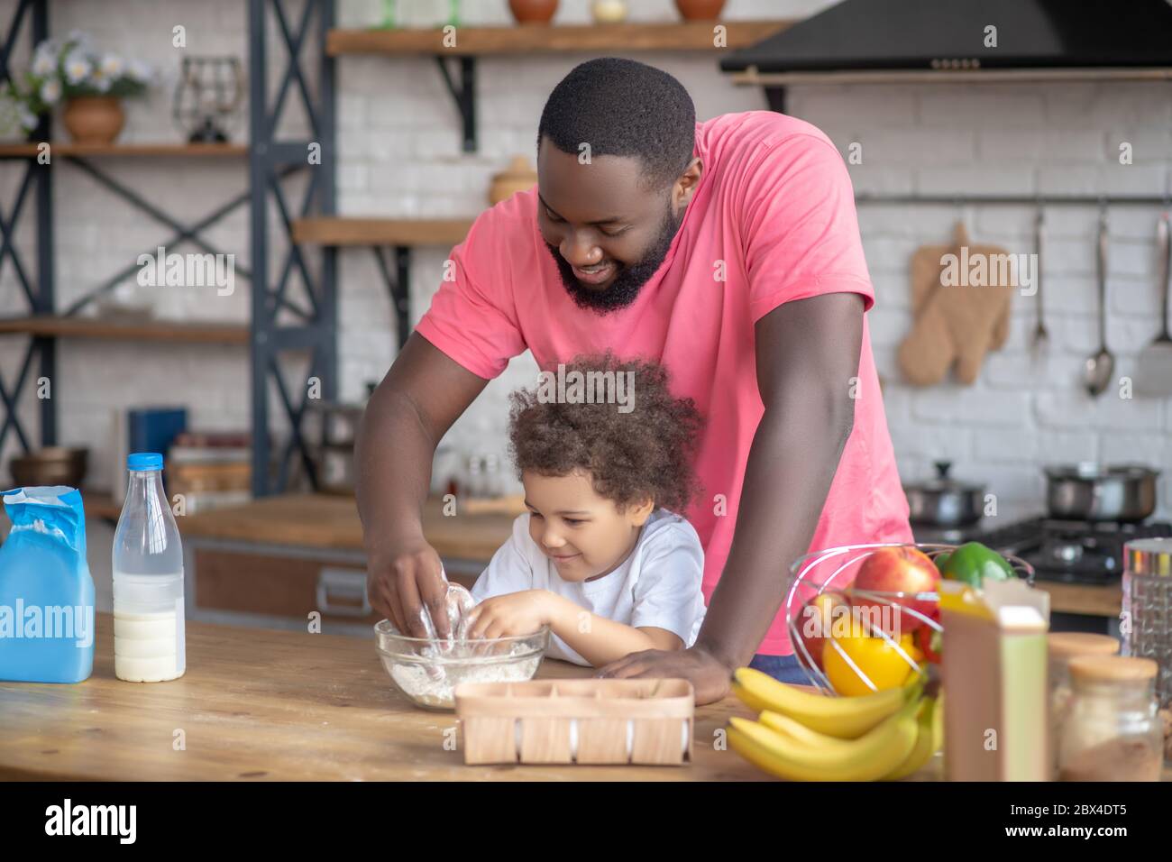 Un hombre de barba afroamericano jugando con su hija removiendo la harina en el cuenco Foto de stock