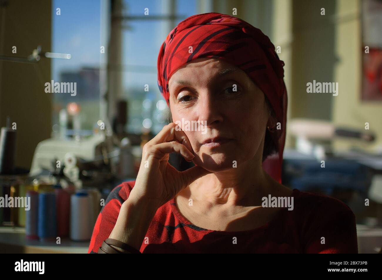 Retrato femenino de mujer madura vestida de ropa roja y sentada al final de su jornada de trabajo en la máquina de coser y el fondo de algodón bobbins Foto de stock