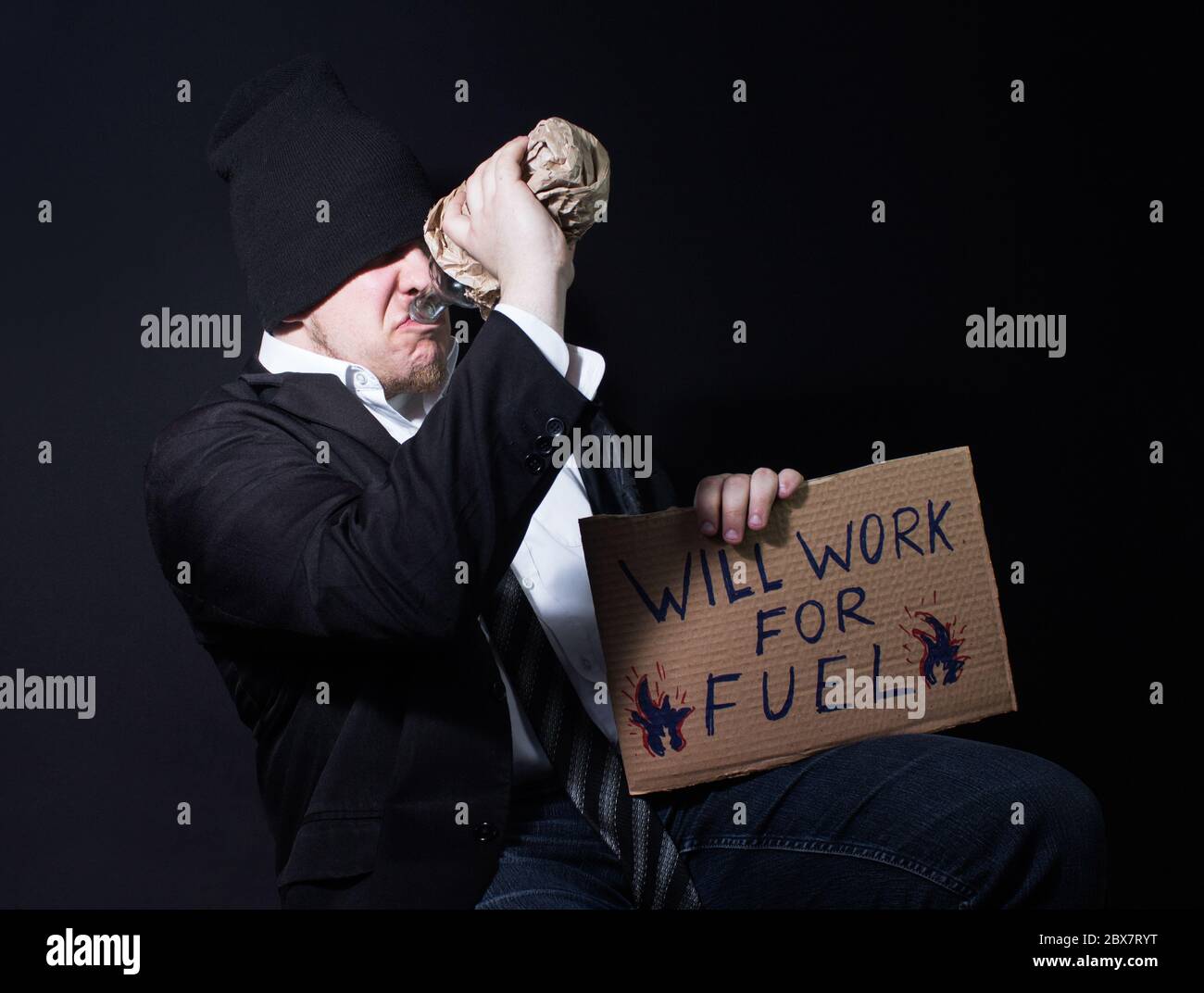 un hombre de negocios sin hogar alchoholic bebiendo de un botín y sentado con el cartón cantar. Foto de stock