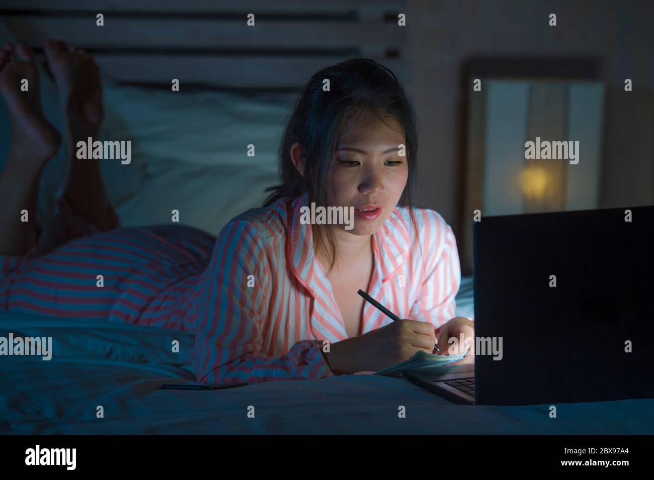 Joven hermosa y segura estudiante de Asia coreana que investiga y estudia en casa preparando el examen con computadora portátil en la cama escribiendo tarde en ni Foto de stock
