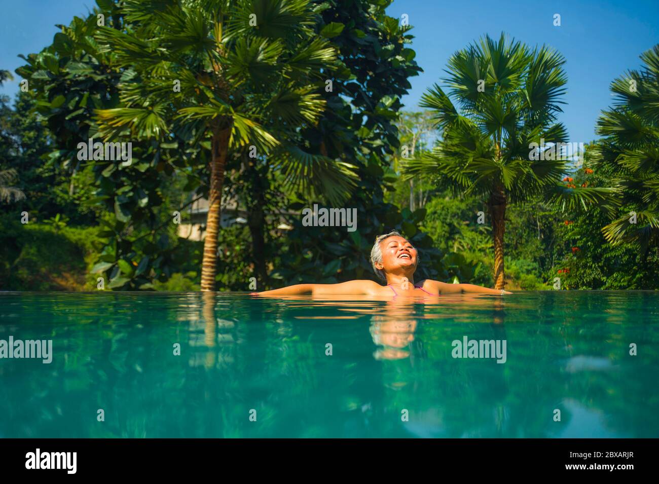 Retrato natural de estilo de vida de una mujer asiática de mediana edad, atractiva y feliz, relajada en un complejo tropical con piscina infinita con fondo de selva Foto de stock