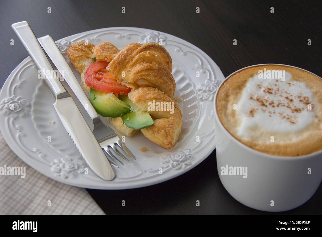 Croissant fresco relleno de aguacate y tomate, una taza de capuchino en una mesa oscura, luz de la mañana desde la ventana Foto de stock