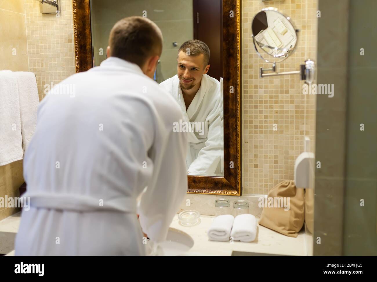 El chico sonríe en un abrigo blanco y se mira en el espejo del baño por la  mañana. Empezar un nuevo día con una sonrisa. Un hombre joven y guapo, de  pie