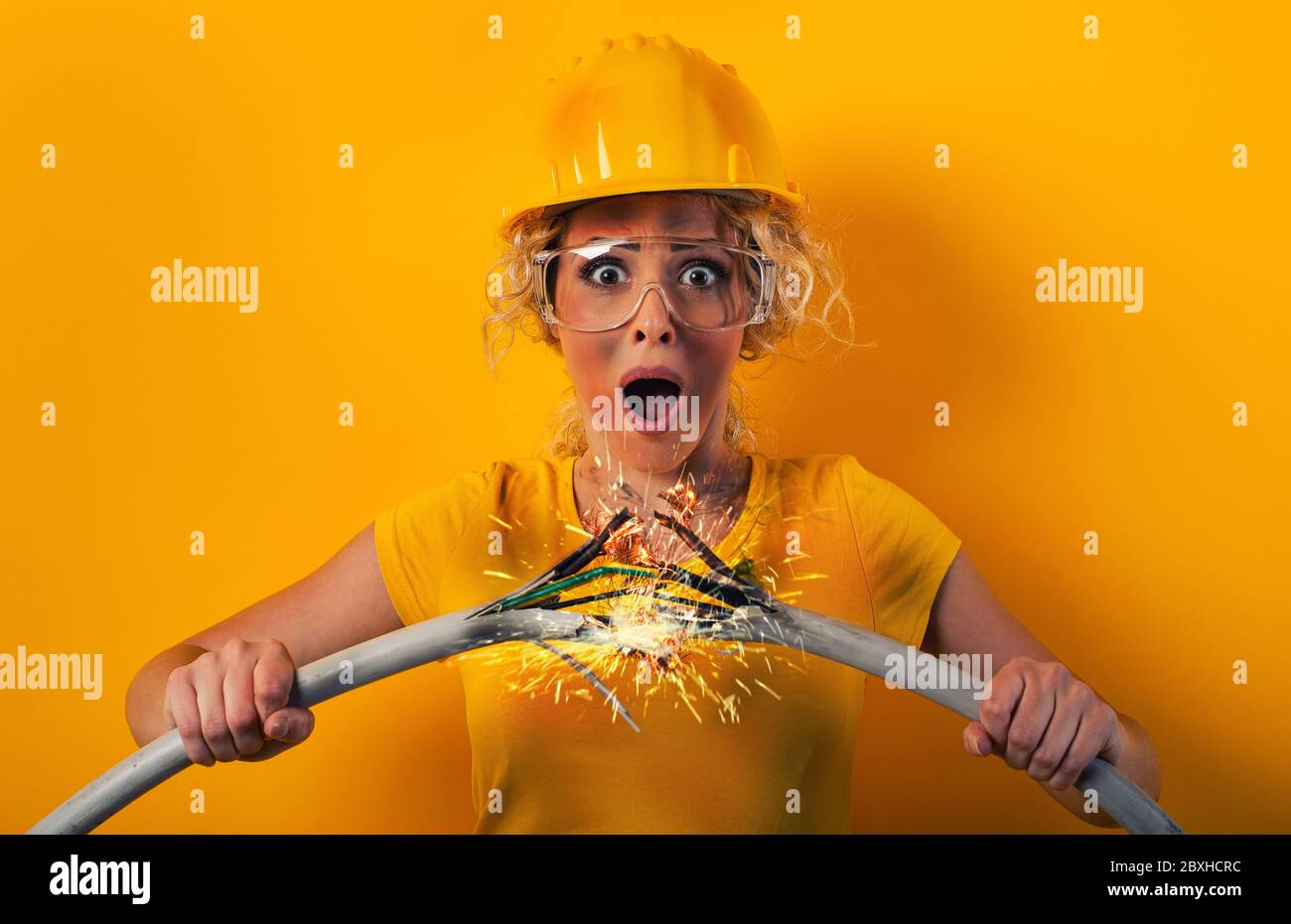 Una chica trabajadora con sombrero rompe un cable eléctrico. Fondo amarillo Foto de stock