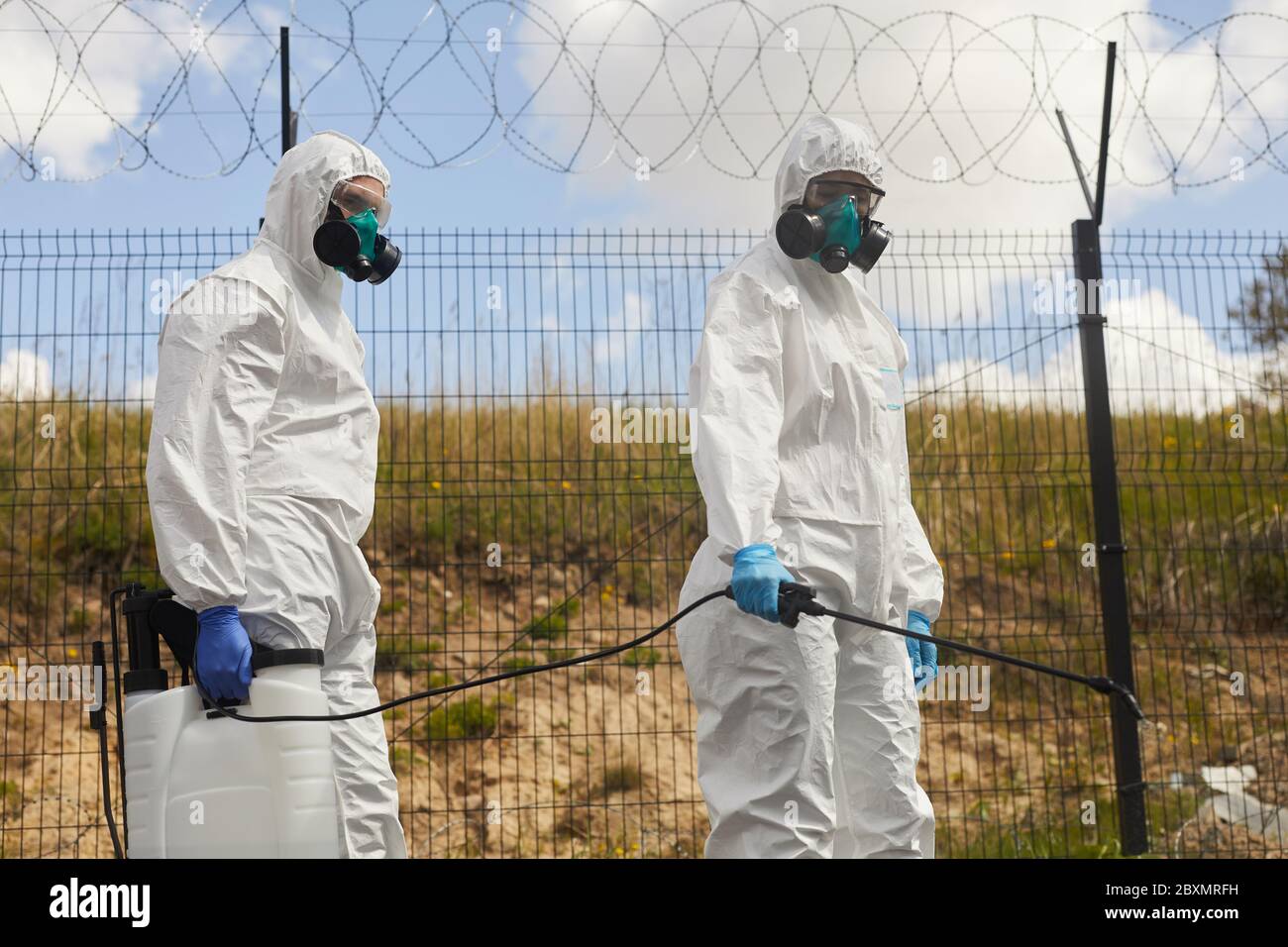 Retrato de dos trabajadores que usan equipo de protección y productos químicos de pulverización al aire libre, parados por una valla de alambre de púas durante la desinfección o limpieza en milígulo Foto de stock