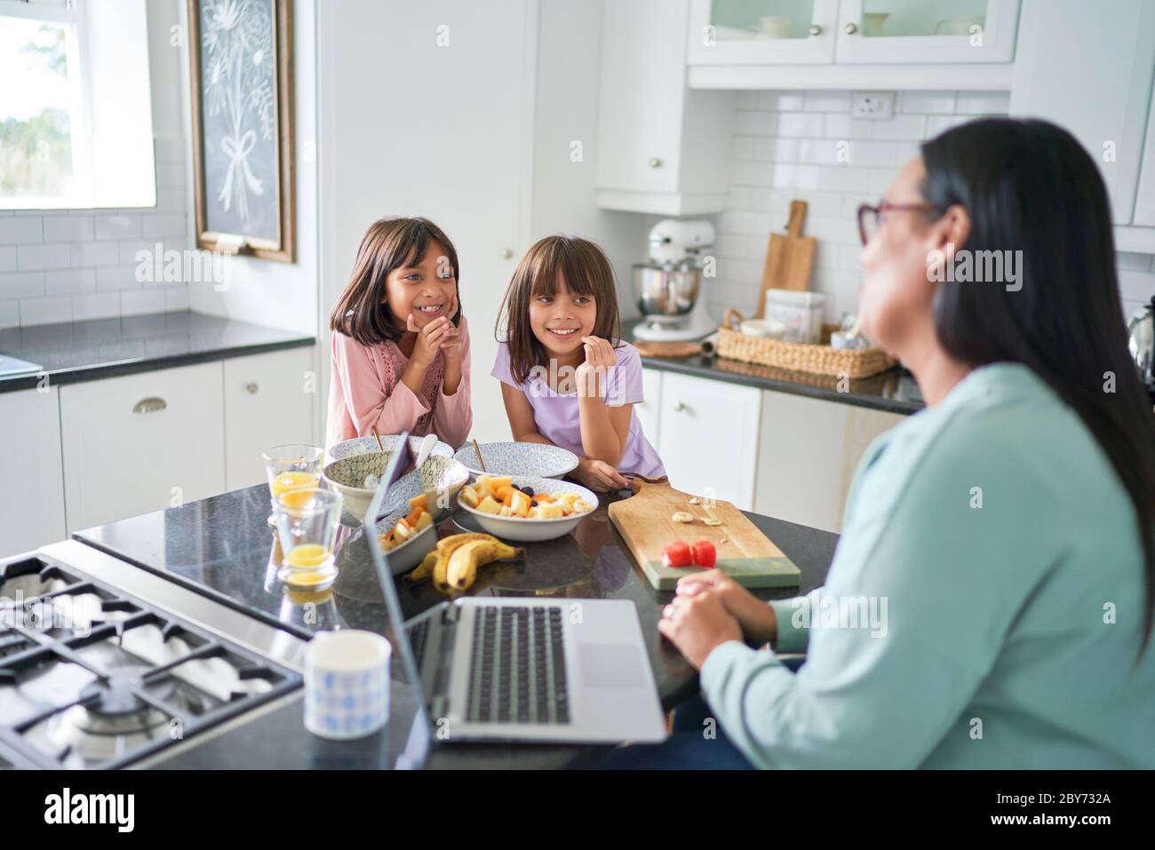 Hijas felices comiendo el desayuno y viendo a la madre trabajar en la cocina Foto de stock
