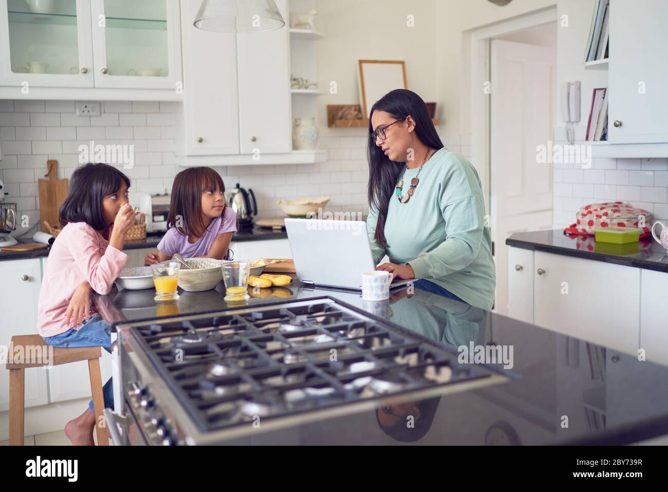 Madre trabajando en el portátil mientras las hijas desayunan en la cocina Foto de stock
