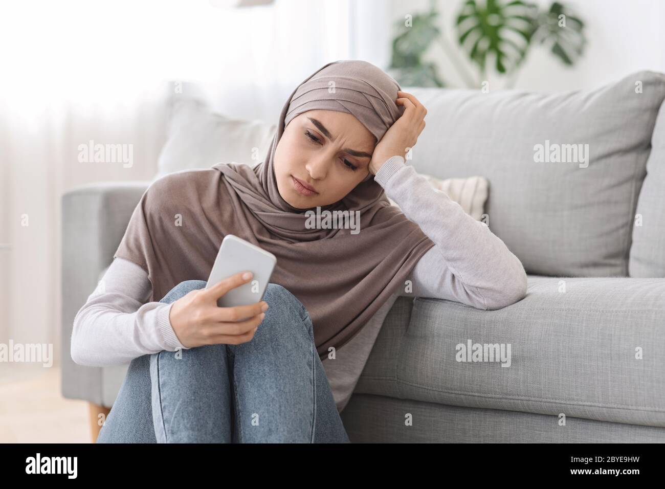 Concepto de depresión. Disgustada mujer árabe sentada con un smartphone en el piso de casa Foto de stock
