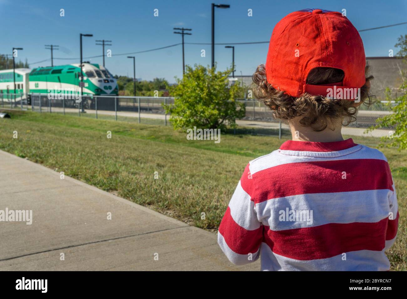 Un niño pequeño con camisa de rayas rojas y gorra roja de béisbol observa mientras un tren se acerca a una estación de ferrocarril Foto de stock