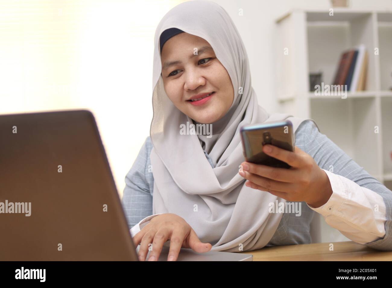 Mujer musulmana asiática trabaja en la oficina, usando el teléfono y el portátil, expresión facial sonriente feliz, compra en línea cheque hacia fuera Foto de stock