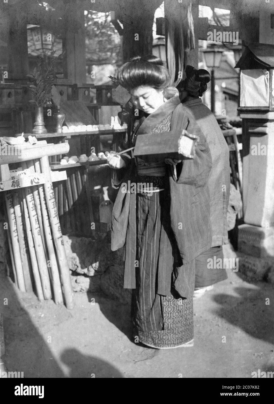 [ 1910s Japón - Mujer japonesa en Shinto Shrine ] — una mujer japonesa está sacando un palo numerado de una caja de madera para contar fortuna en un Shinto Shrine. El número del palo hace referencia a un pedazo de papel numerado, llamado o-mikuji, con descripciones de la fortuna, salud, relaciones románticas y otros aspectos de la vida diaria. En las puertas miniatura torii sagrado el año Taisho 5 (1916) está escrito y el 1 de enero. Así que esta mujer está visitando el santuario durante el hatsumode (初詣), la primera visita Shinto santuario del año Nuevo Japonés. siglo 20 vintage gelatina de plata. Foto de stock