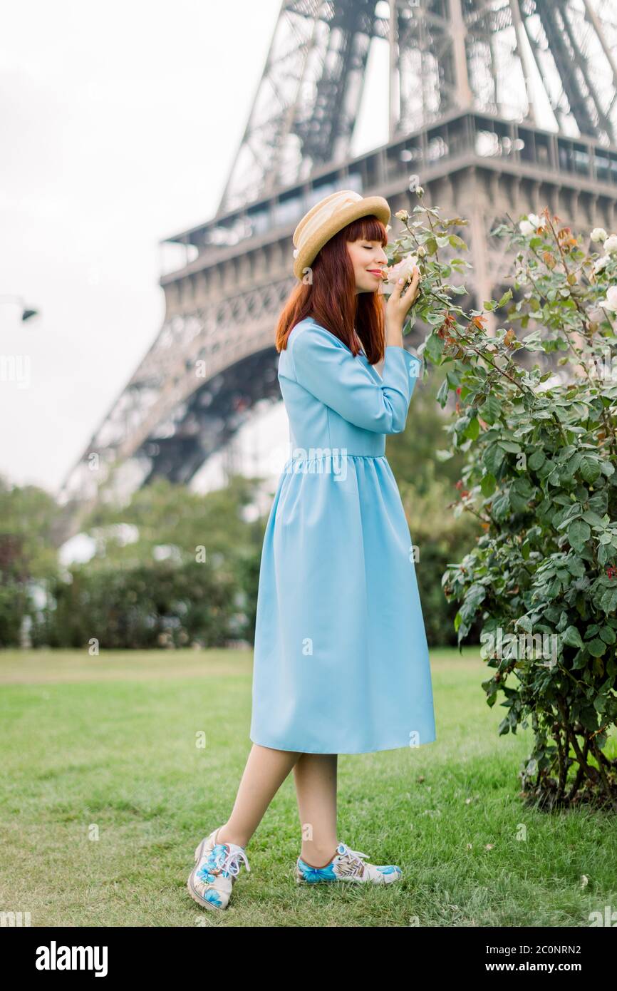 Joven hermosa mujer, vestido con elegante vestido azul claro y sombrero de  paja, camina por la calle de la ciudad de París cerca de los rosales  florecientes, tocar y Fotografía de stock -