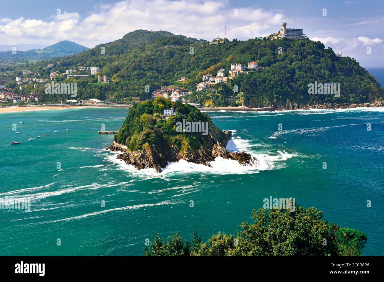 Vista a la pequeña isla verde en medio del océano verde y la colina con el edificio en la parte superior rodeado de playa de arena (San Sebastián) Foto de stock