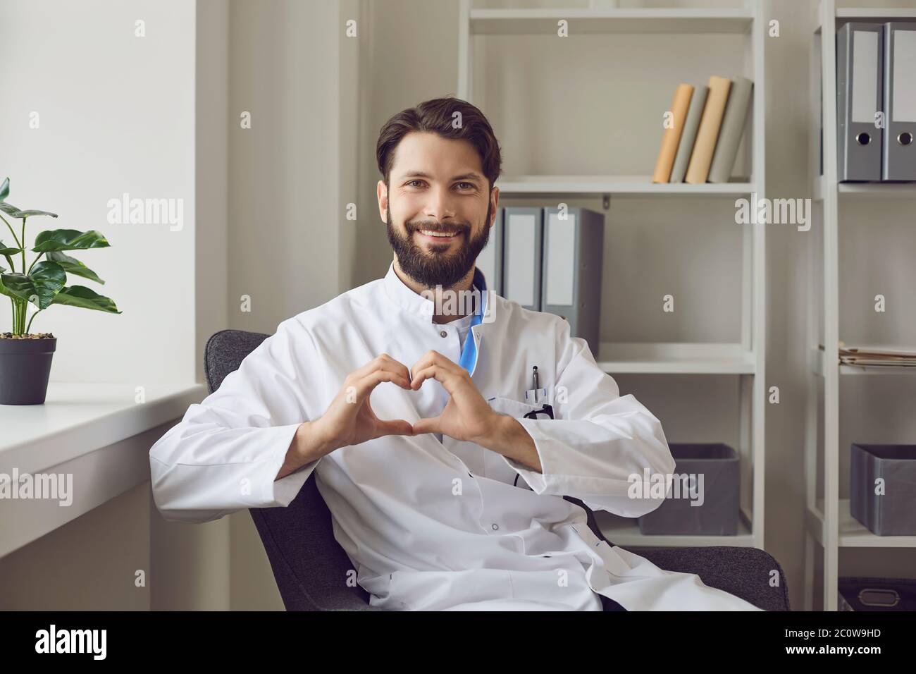 Feliz médico joven mostrando un gesto de corazón en su oficina. Salud del corazón, donación o caridad. Atención médica sobre los pacientes Foto de stock