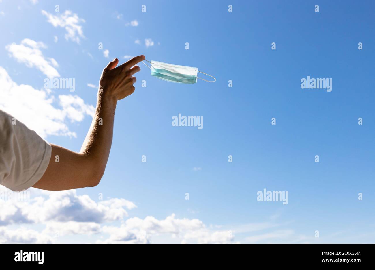 Fin de cuarentena, covid-19. El hombre que quita y tira la máscara médica protectora usada, la sostiene en su dedo sobre el fondo azul del cielo, goza de la vida, b Foto de stock