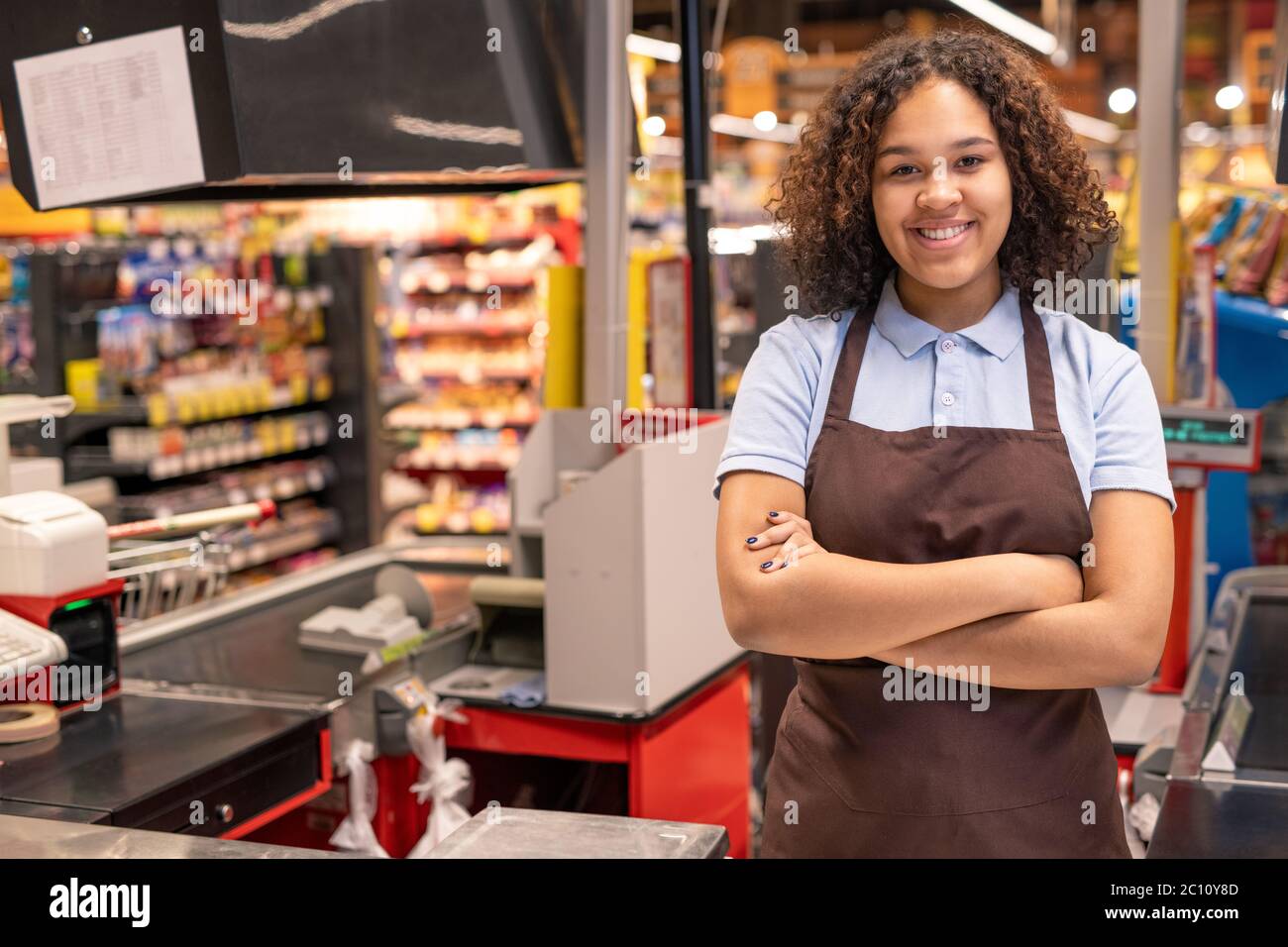 Joven asistente de tienda o cajero sonriente en el trabajo de cruzar los  brazos por el pecho mientras que de pie por el lugar de trabajo en el  entorno de supermercado Fotografía