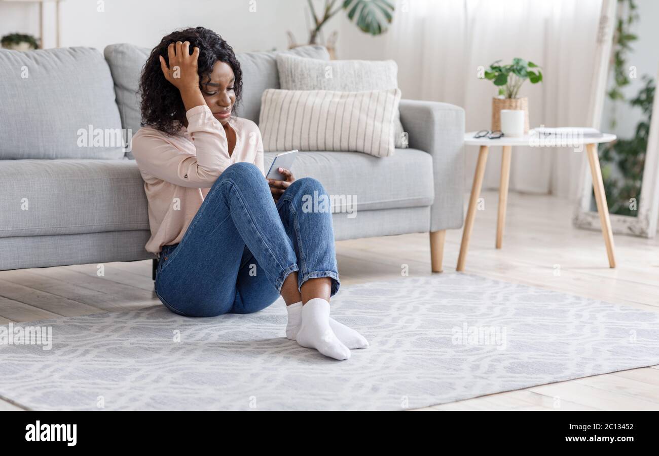 Chica desgarrada. Llorando Mujer Negra sentada con un smartphone en el piso de casa Foto de stock