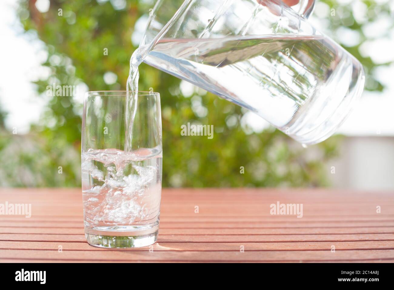 Agua potable que fluye de una jarra de cristal a un cristal, de fondo con plantas Foto de stock