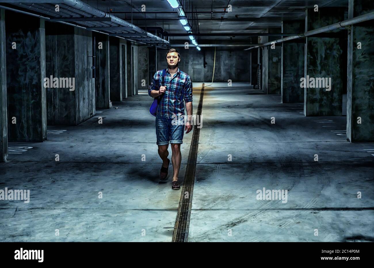 Un joven caminando por un sótano oscuro en un edificio comercial a lo largo  del canal de drenaje llevando una bolsa sobre su hombro acercándose a la  cámara Fotografía de stock -