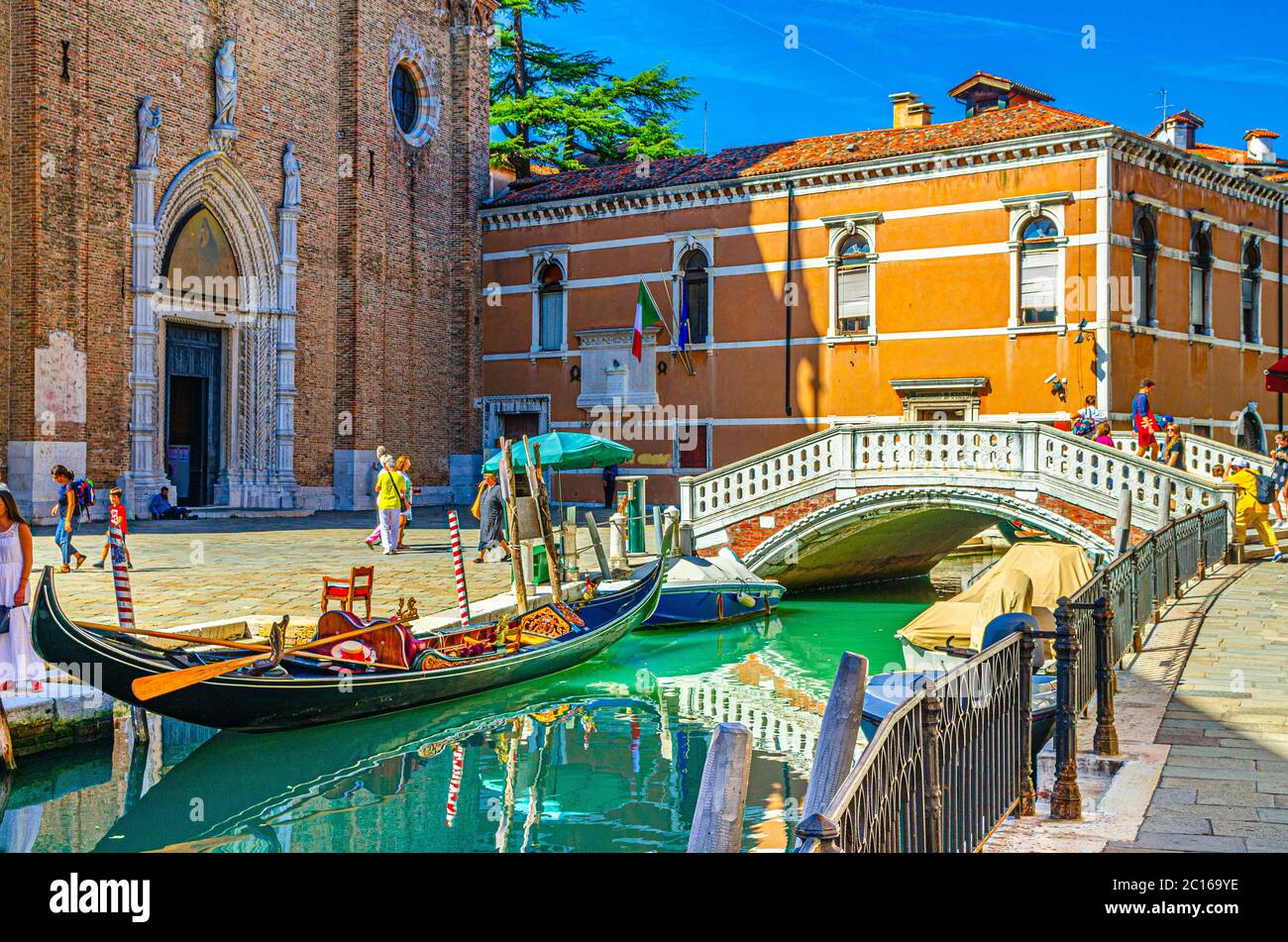 Venecia, Italia, 13 de septiembre de 2019: Góndolas y barcos amarrados en el estrecho canal de agua, Basílica di Santa Maria Gloriosa dei Frari Iglesia católica y puente de piedra, Región Veneto, cielo azul en el día de verano Foto de stock