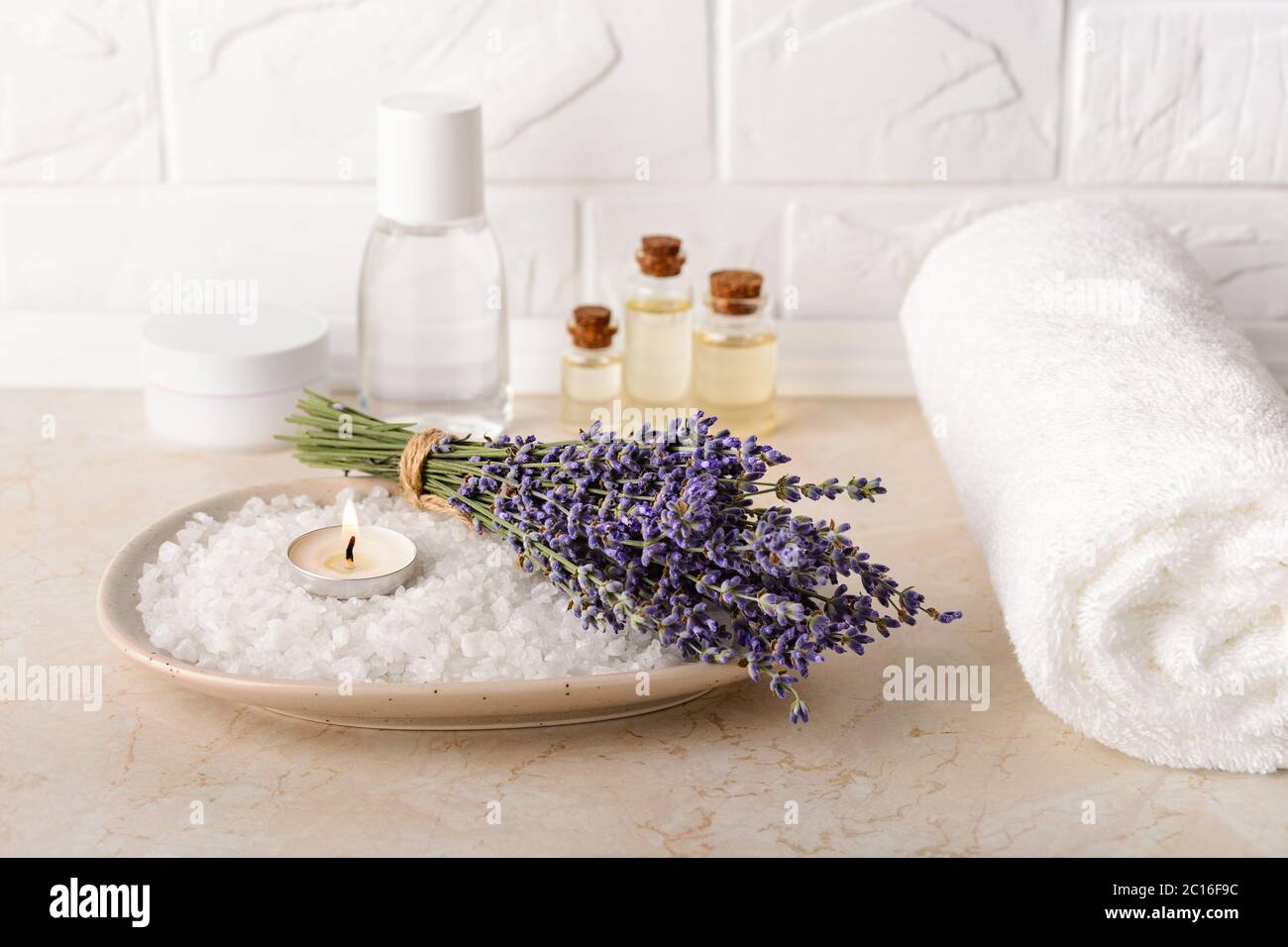 Lavanda fresca y aromática vela ardiente en una sal de baño cerca de toalla  blanca de felpa en un baño. Spa casero, cuidado de la piel y cosmetología  Fotografía de stock -