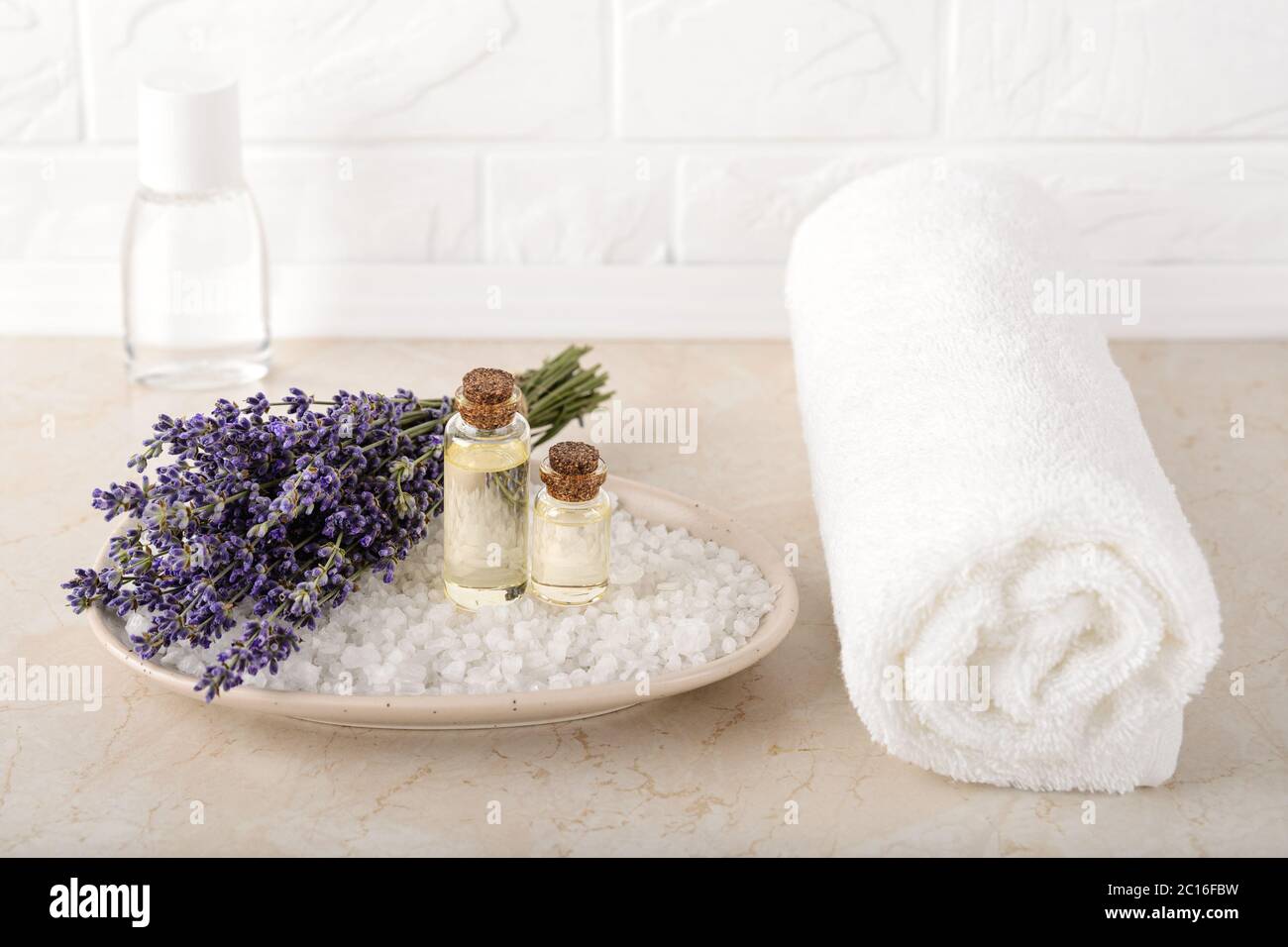 Lavanda fresca y aromática aceite esencial en una sal de baño cosmética  cerca de toalla blanca de felpa en un baño. Spa casero, cuidado de la piel  y cosmetología Fotografía de stock -
