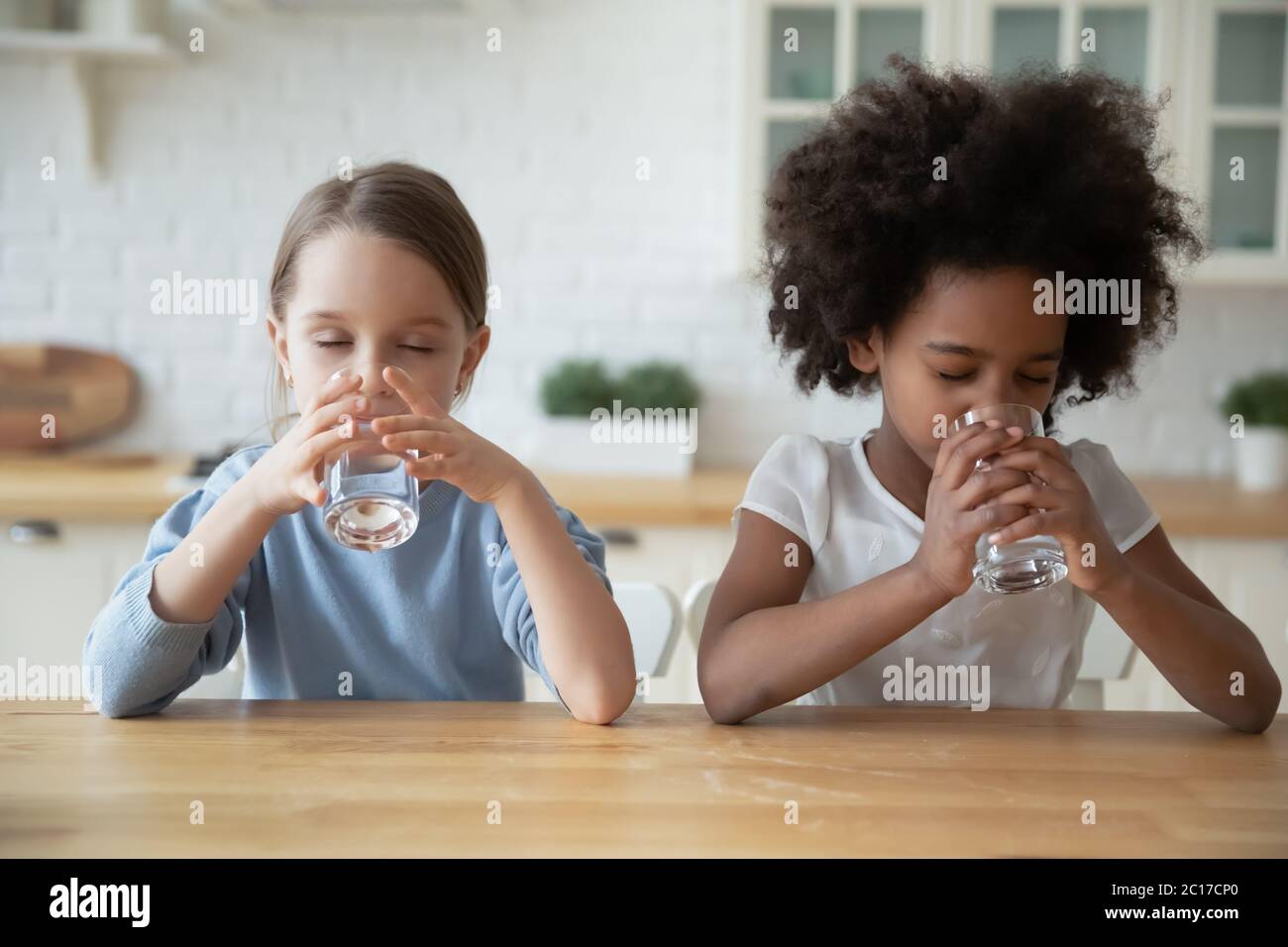Dos niñas multiétnicas que beben agua natural sentadas en la cocina Foto de stock