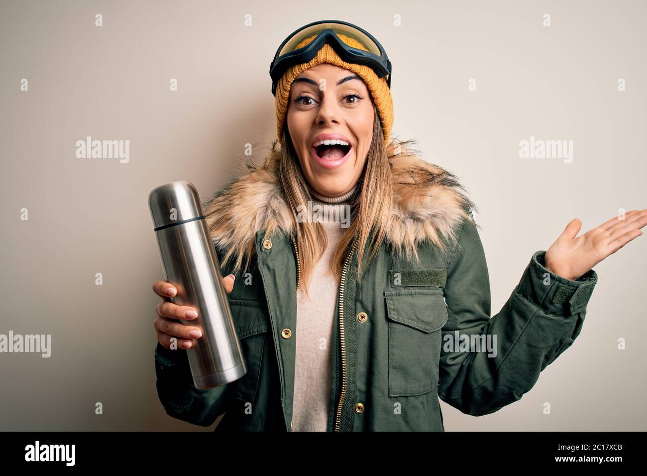 Joven hermosa mujer mosa morena con gafas de esquí tomando termo con café muy feliz y emocionado, la expresión ganadora celebrando la victoria scre Foto de stock