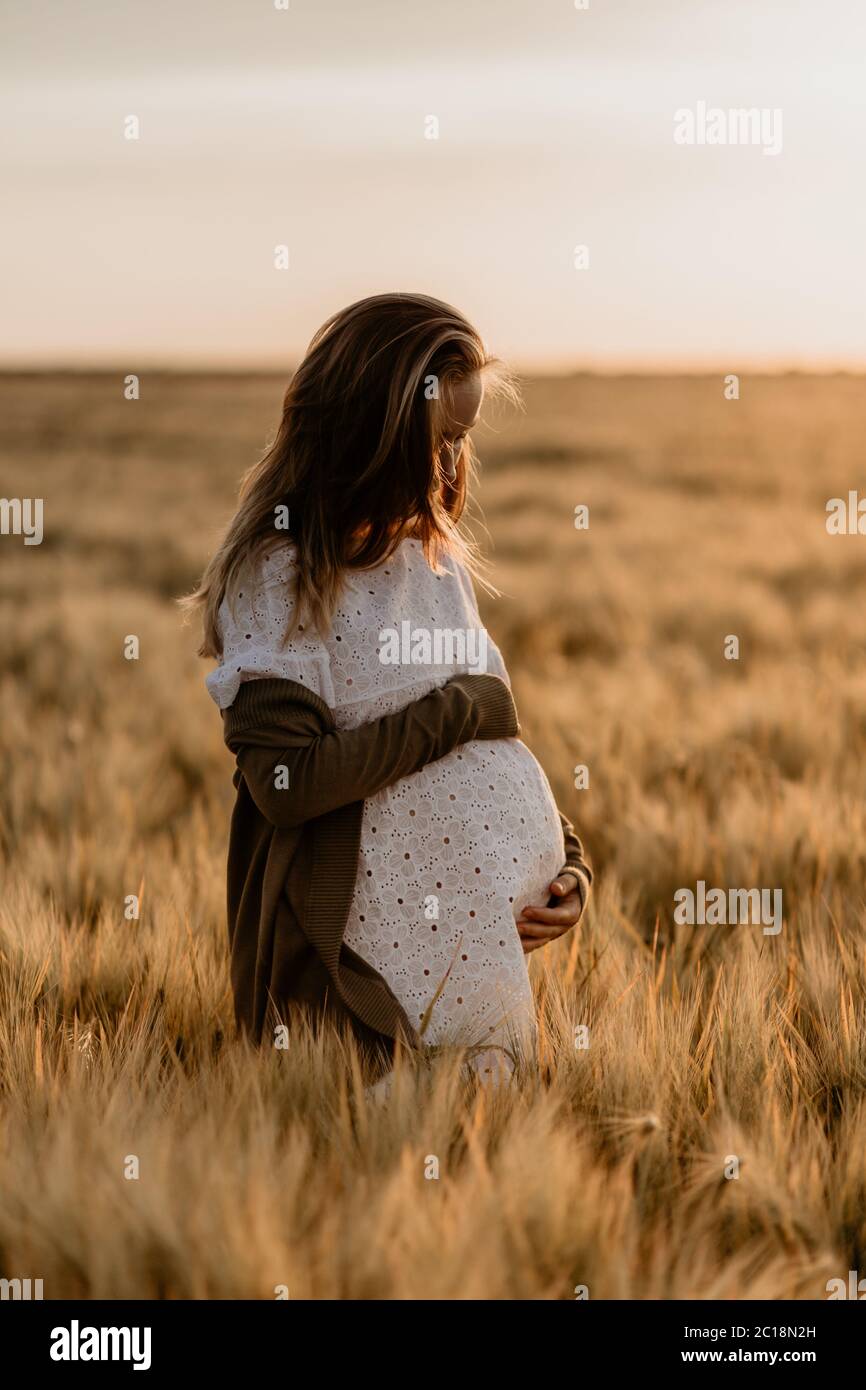 Joven hermosa mujer embarazada mirando hacia abajo y tocando su vientre en el campo de trigo naranja en un soleado día de verano. La naturaleza en el país. Milagro Foto de stock
