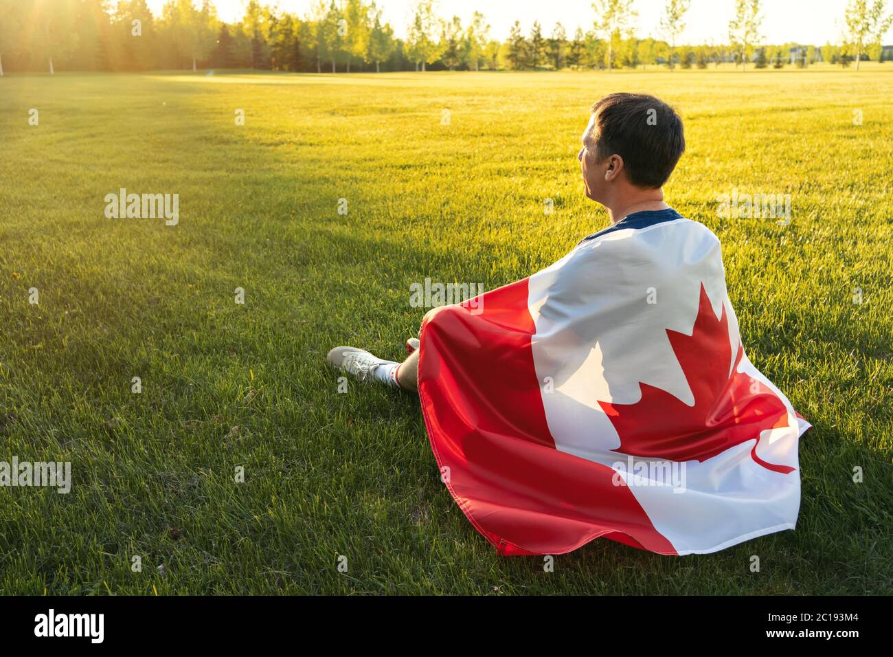 Concepto de día de Canadá. Los hombres de edad media están sentados en la hierba en el parque con la bandera canadiense en su espalda. Concepto de vacaciones nacionales Foto de stock