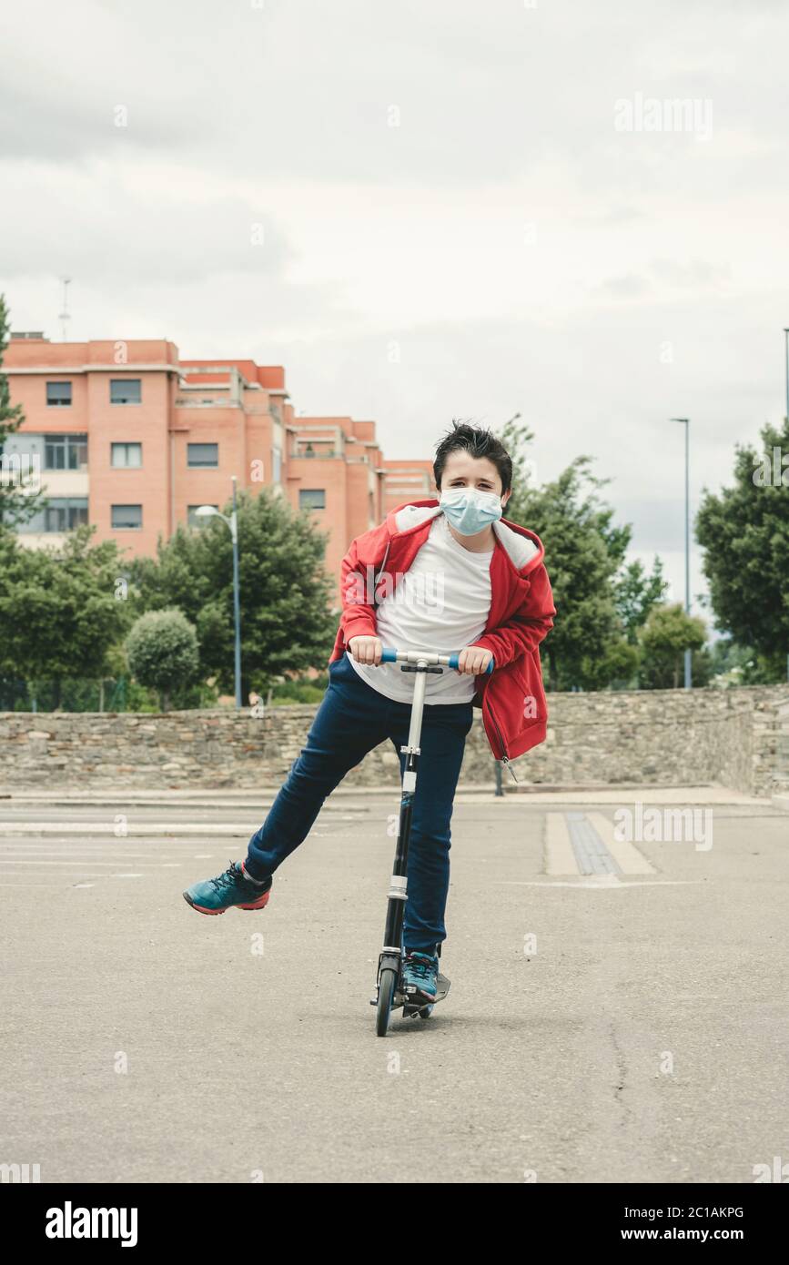 niño feliz usando máscara médica con su scooter en la ciudad al aire libre Foto de stock