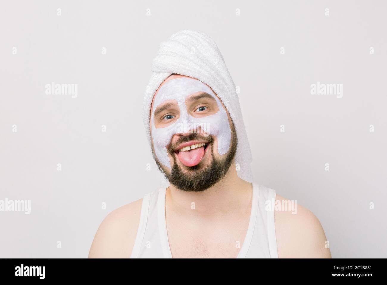 Joven loco barbudo en toalla blanca, con máscara facial blanca en la cara.  Foto de hombre con piel perfecta, sonriendo y mostrando lengua. Belleza y  Fotografía de stock - Alamy