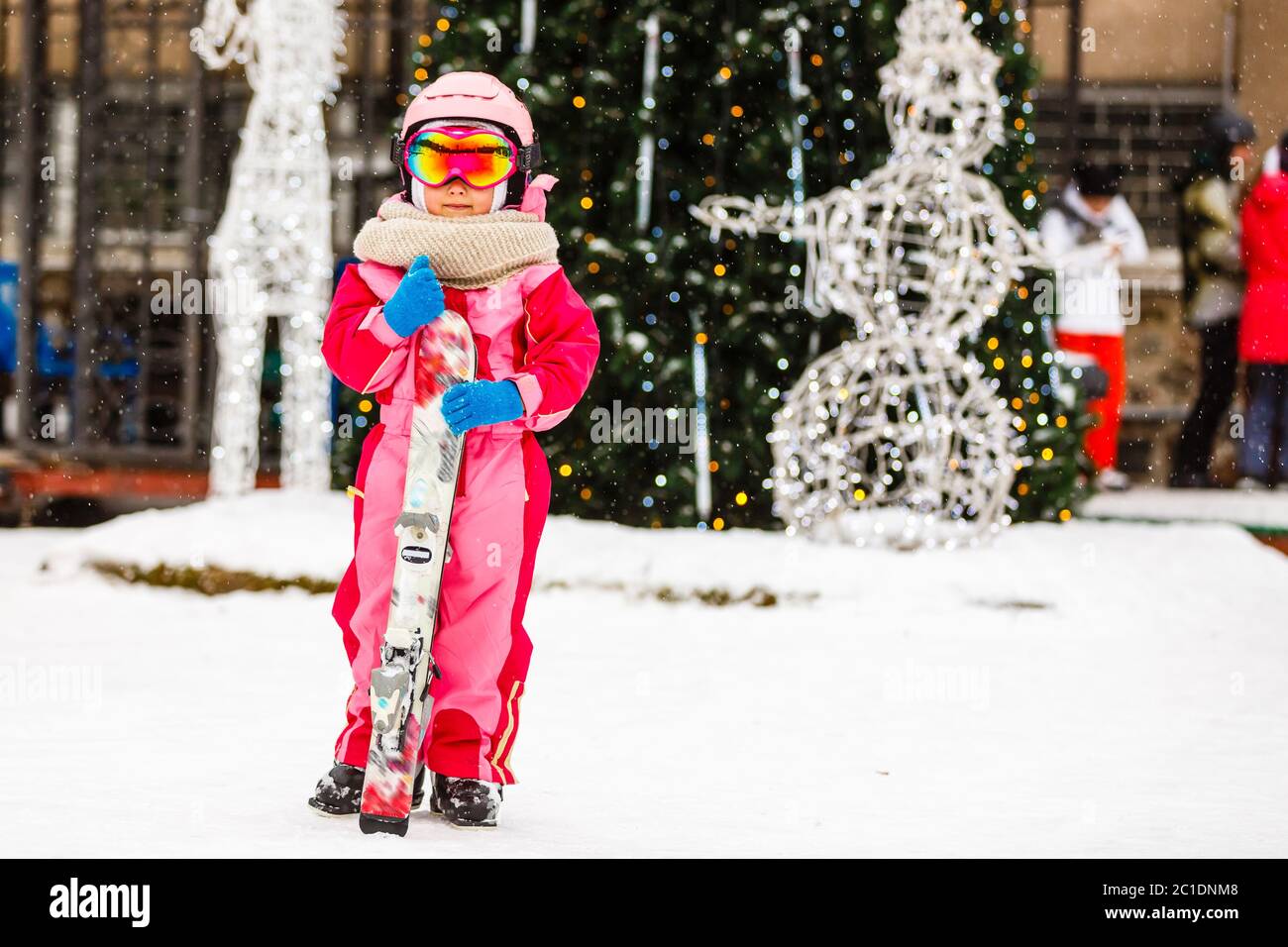 Niña gafas protectoras y un casco en vacaciones de invierno al aire libre fijación de esquís detrás buscando cámara sonriendo emocionada cerca fondo borroso Foto de stock