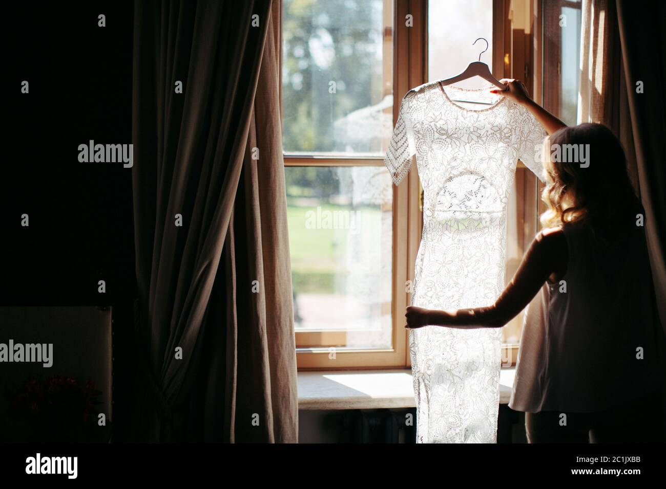 Mujer rubia con pelo rizado sosteniendo y mirando el hermoso vestido blanco de boda en un perchero, de pie cerca de la ventana en su casa, vista de atrás, daylig Foto de stock