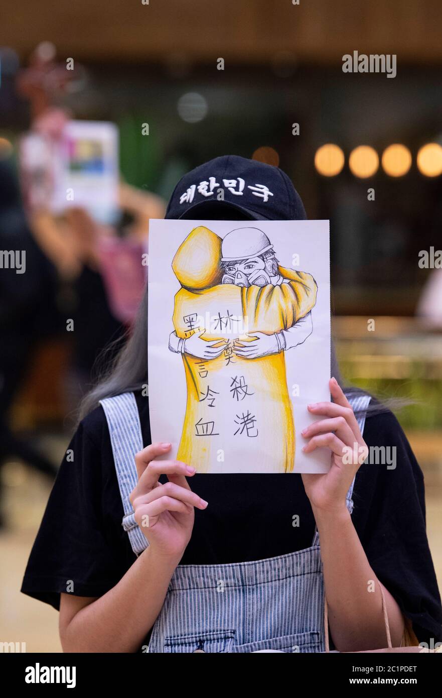 HONG KONG,HONG KONG RAE,CHINA: 15 DE JUNIO DE 2020. Un manifestante sostiene una obra de arte que muestra al “hombre impermeable” en el centro comercial Pacific Place Mall en Admiralty Hong Kong, mientras que otros asoman a favor de la independencia y firman “Gloria a Hong Kong”; el himno de protesta. Están allí para conmemorar la muerte de los primeros manifestantes democráticos hace un año. Marco Leung cayó a su muerte por andamios fuera del edificio el 15 de junio de 2019. Se le conoció como el hombre impermeable. Imagen de stock de Alamy/Jayne Russell Foto de stock