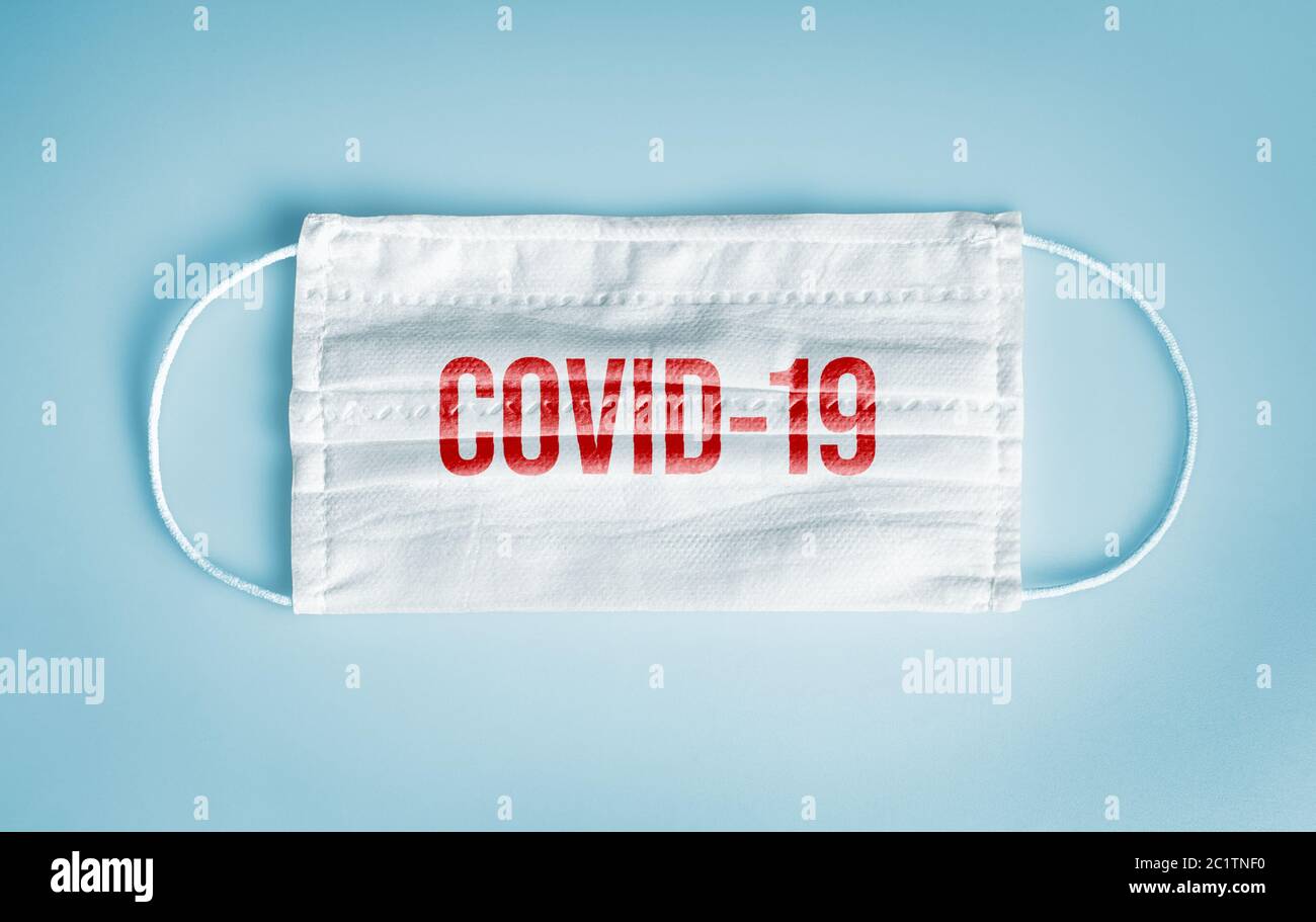 Covid-19 concepto de protección contra el coronavirus y distanciamiento social: Mascarilla facial desechable médica con mensaje covid-19 sobre fondo azul. Foto de stock