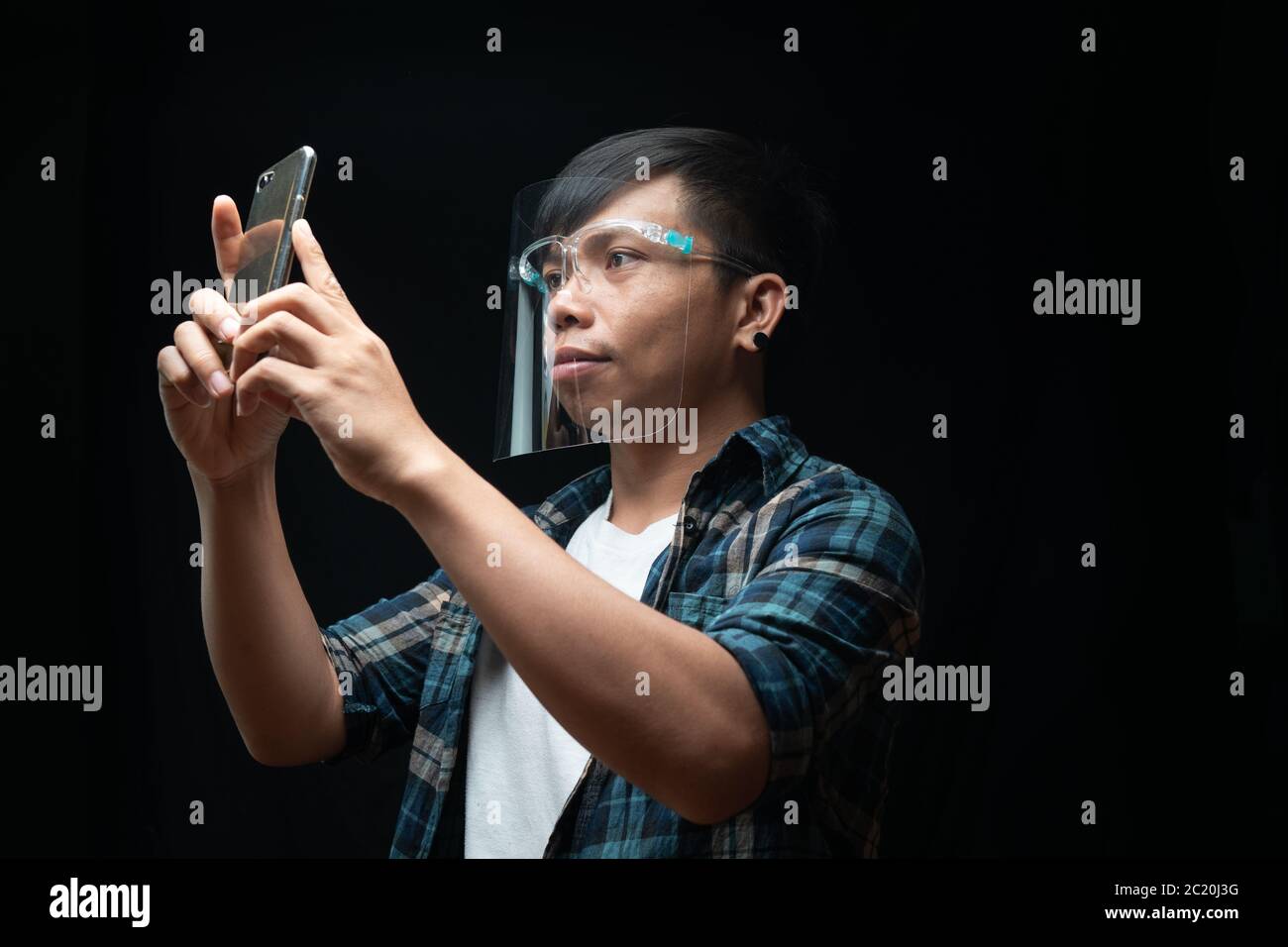 El hombre lleva un protector facial utilizando el smartphone hacer una foto sobre el fondo negro aislado Foto de stock