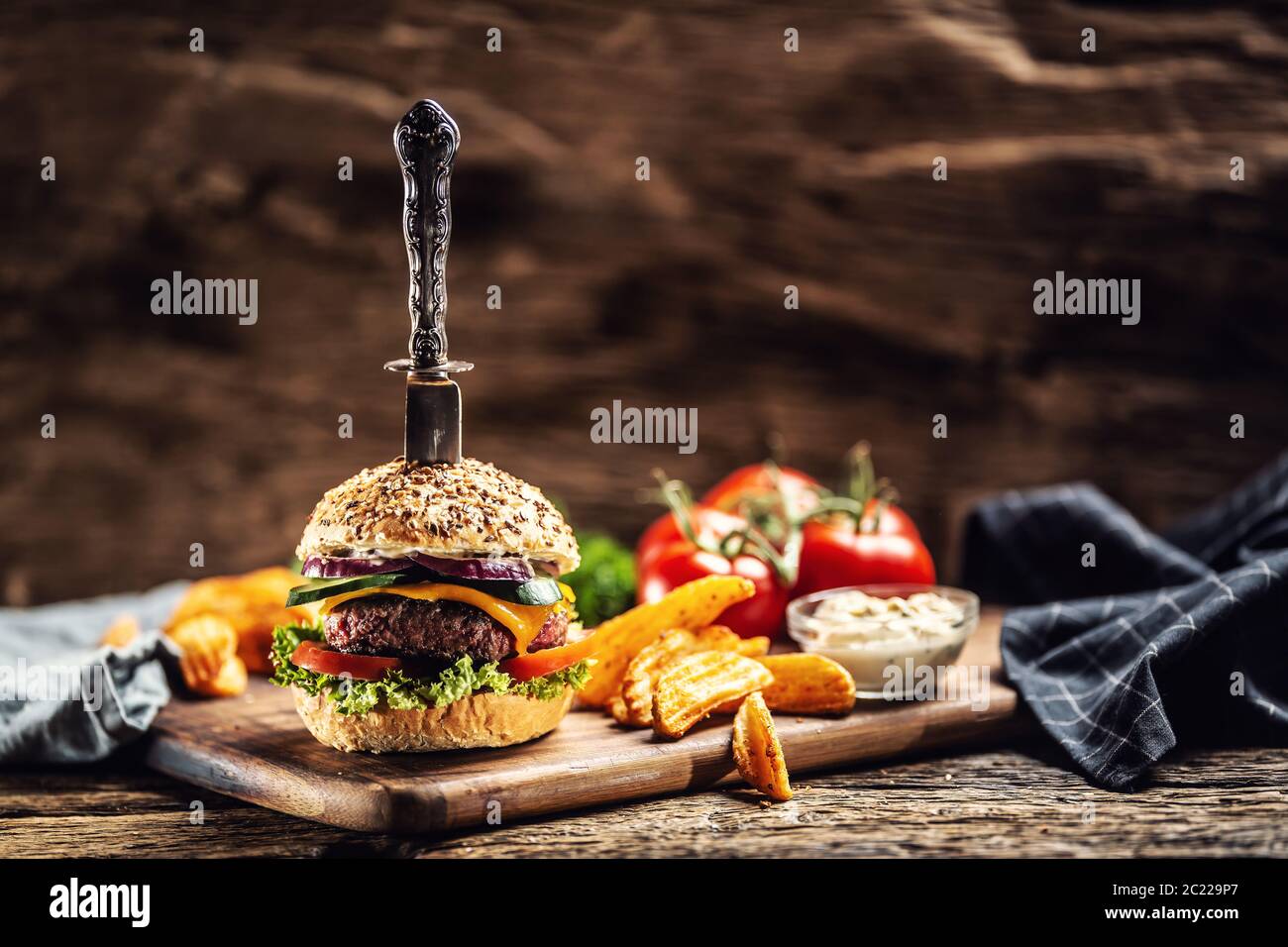 Cuchillo apuñalado en una hamburguesa con cuñas de patata en un lado en un ambiente rústico oscuro Foto de stock