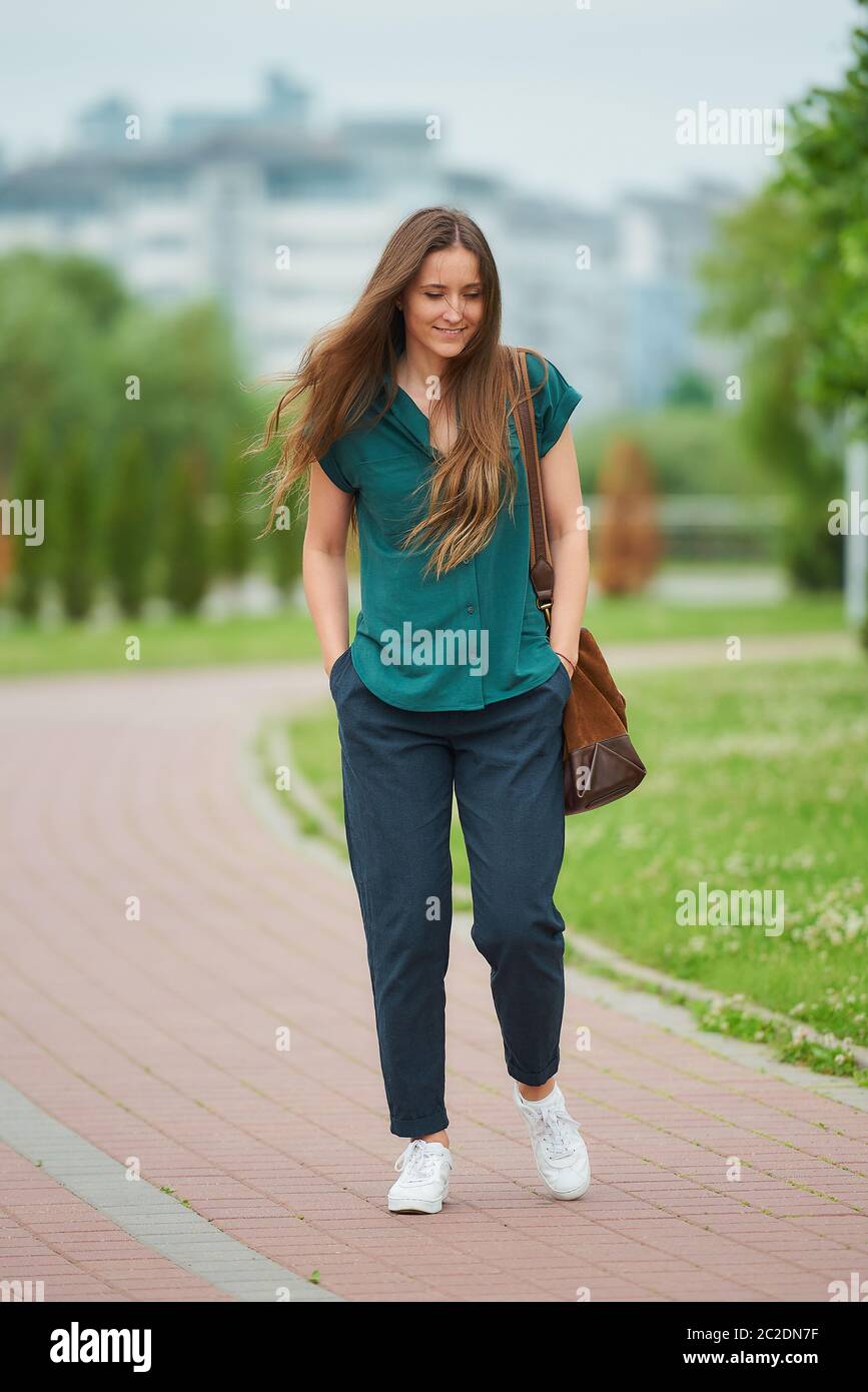 Una mujer lleva ropa elegante y casual disfrutando de su caminar con las manos en los bolsillos de los pantalones en el parque. Una chica bonita con pelo Fotografía de stock -