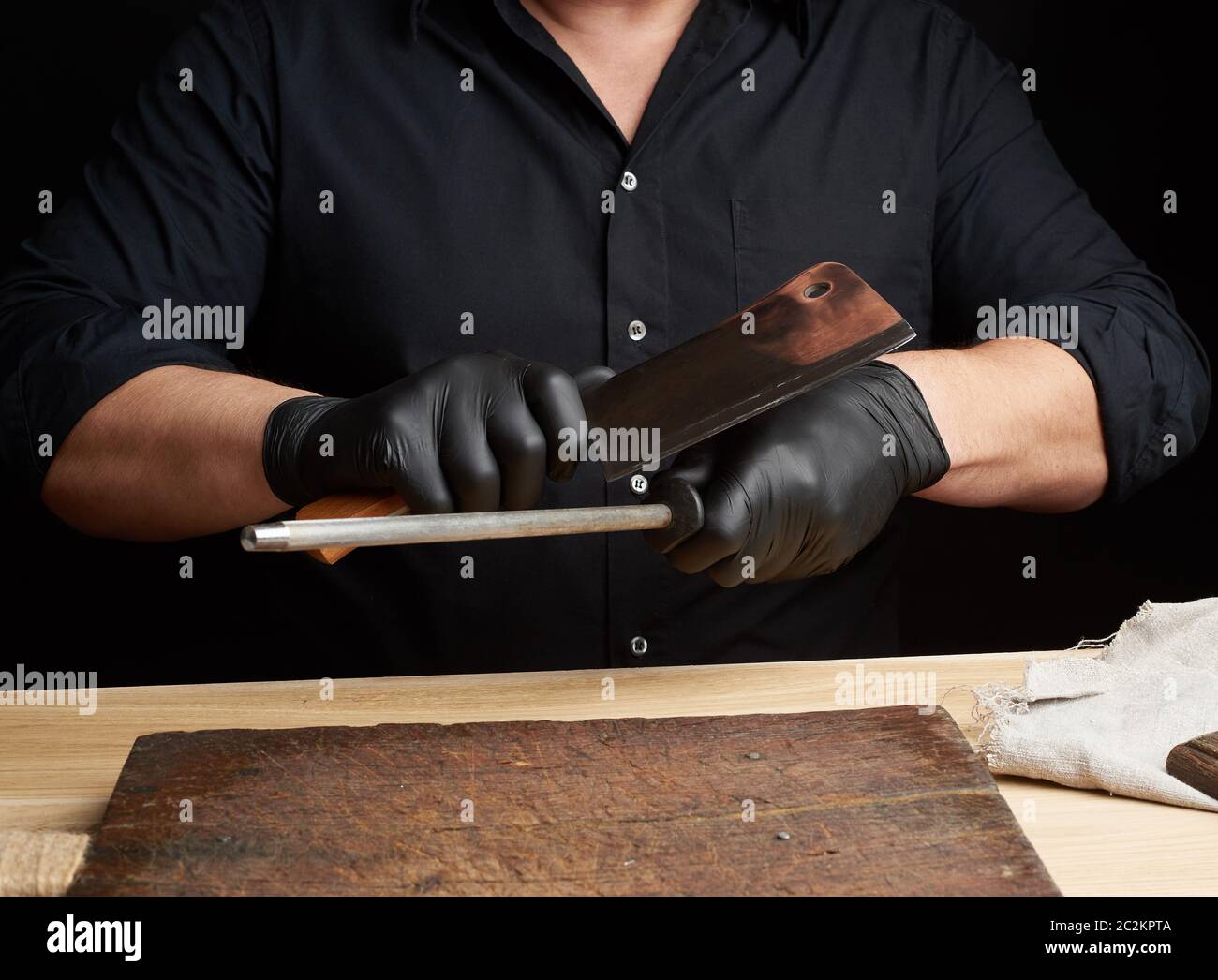 Chef en una camisa negra y guantes de látex negro afilar un cuchillo de  cocina en un afilador de hierro con un mango encima de la tabla, bajo  llave, cerrar Fotografía de