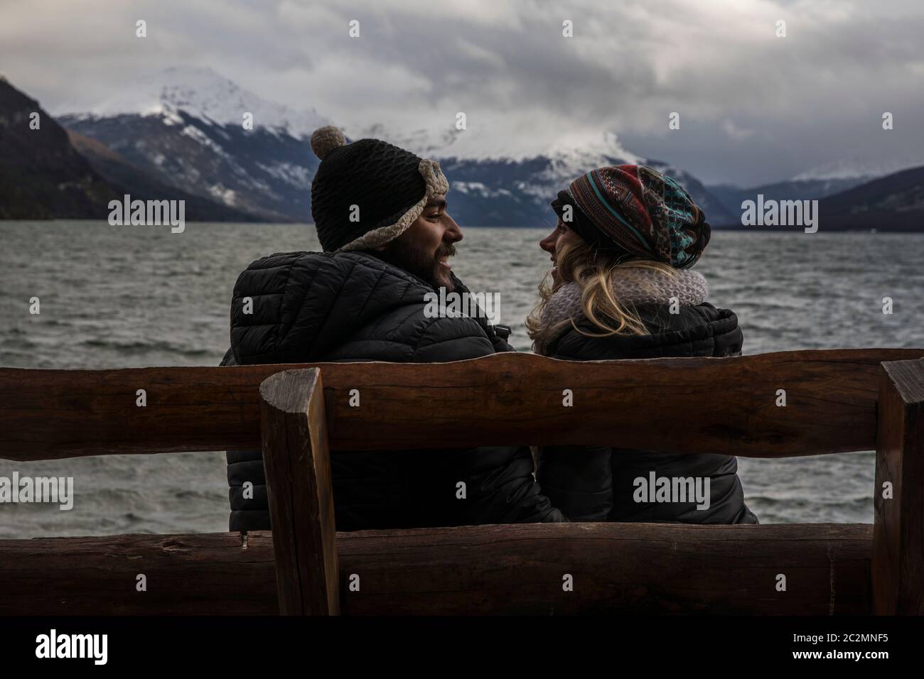 feliz pareja sentada en un banco en la playa frente al océano con una montaña con nieve en la cima en el fondo Foto de stock